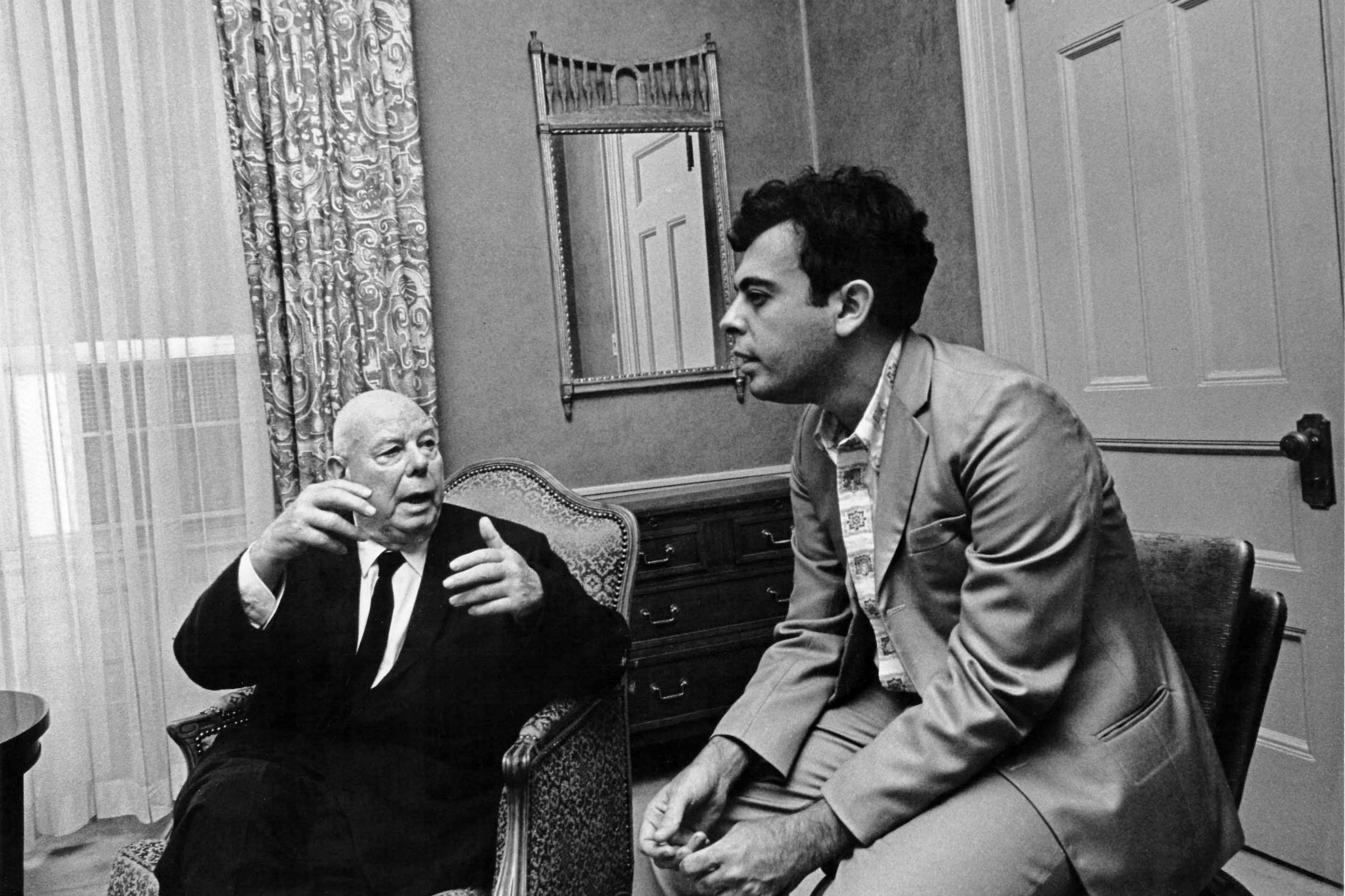 Dois homens sentados em cadeiras em uma sala. Homem mais velho à esquerda, de terno escuro, gesticula com as mãos enquanto fala. Homem mais jovem à direita, vestido com terno claro, escuta atentamente. Ao fundo, cortina estampada, espelho na parede e porta fechada.