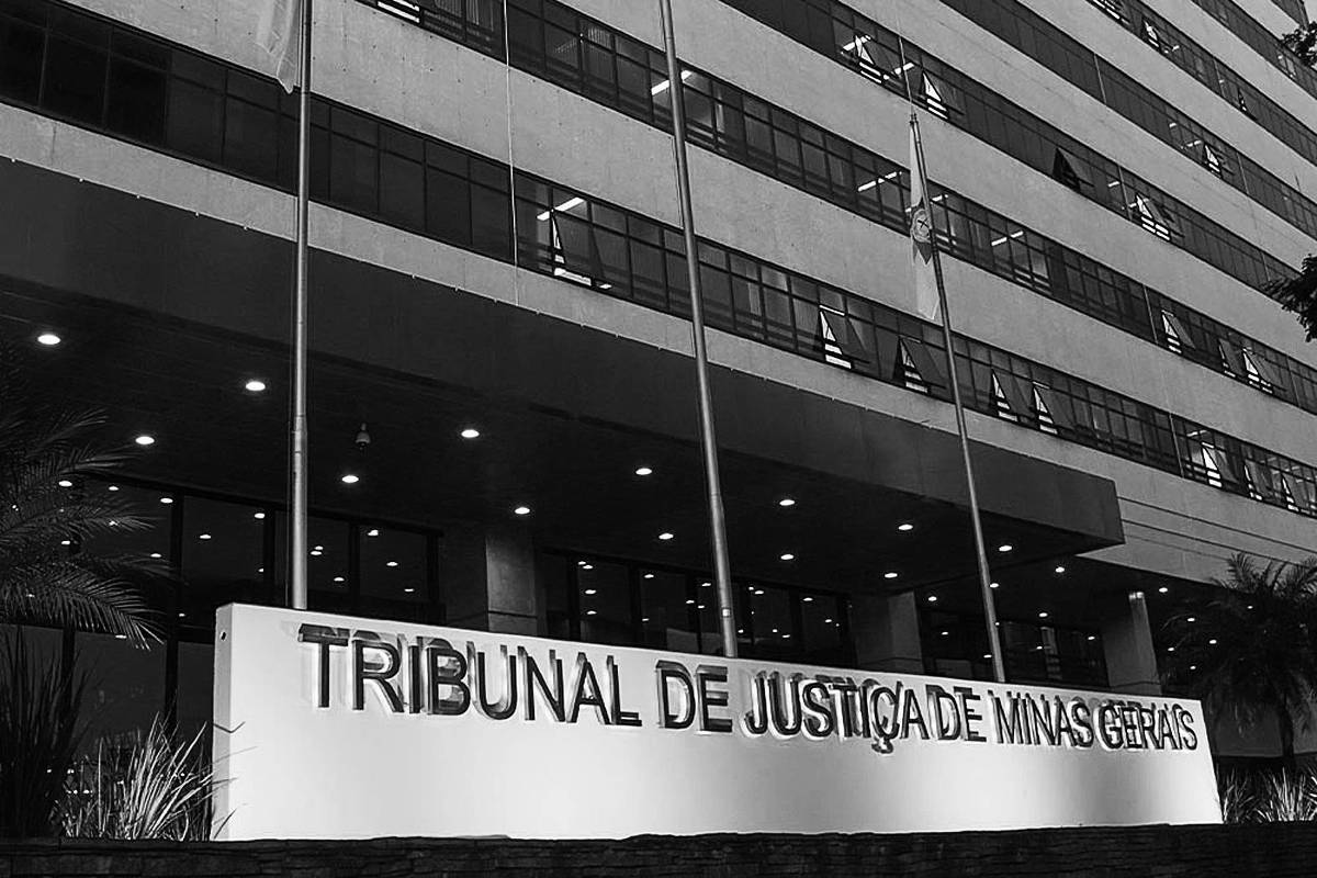 Fachada iluminada do Tribunal de Justiça de Minas Gerais com letreiro em destaque na frente. Prédio grande com várias janelas e bandeiras em mastros visíveis.