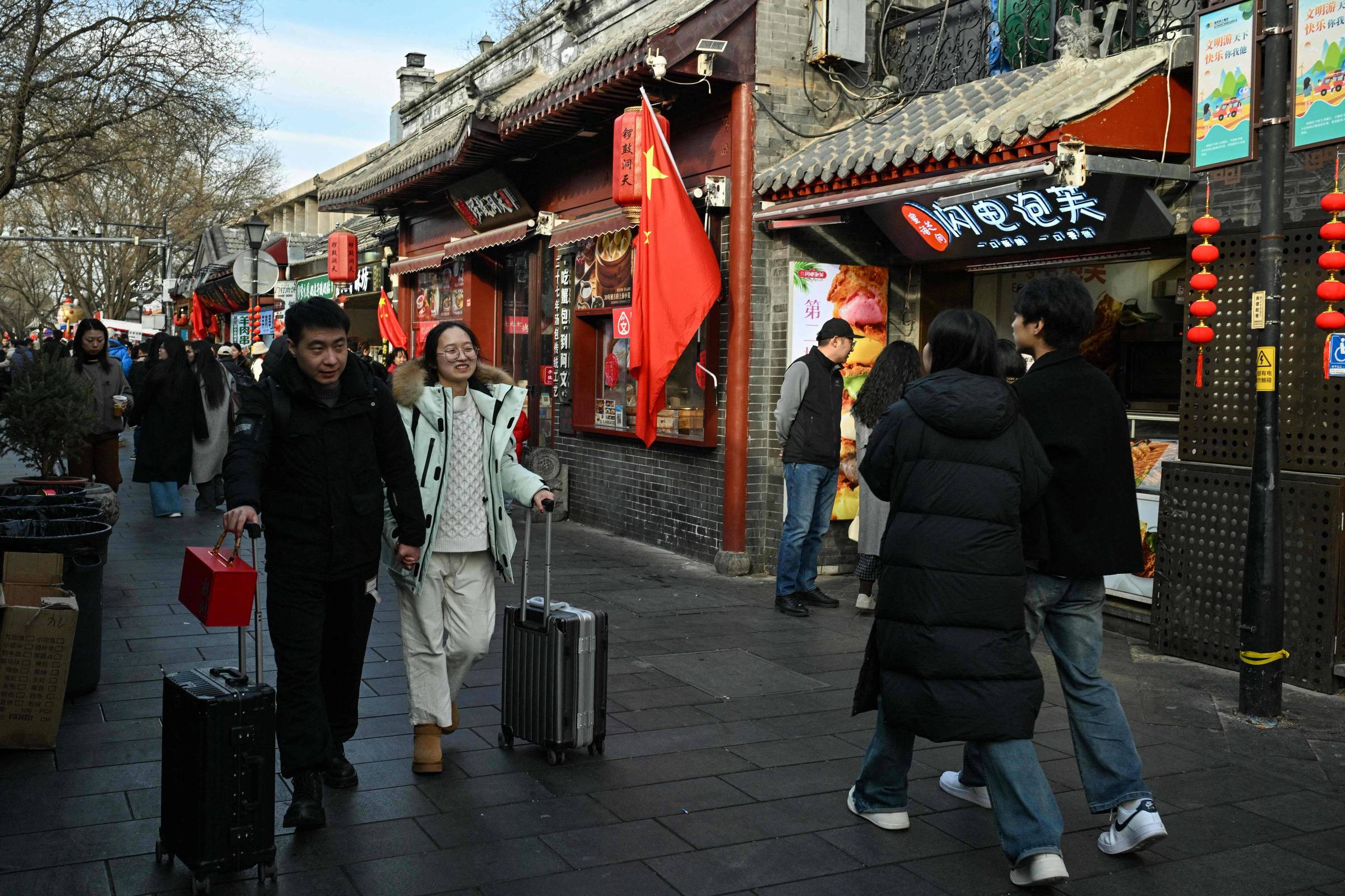 Pessoas caminham por calçada em rua com arquitetura tradicional chinesa e lanternas vermelhas. Duas pessoas puxam malas, uma delas segura bandeira da China. Fachadas exibem letreiros em chinês e decoração típica.