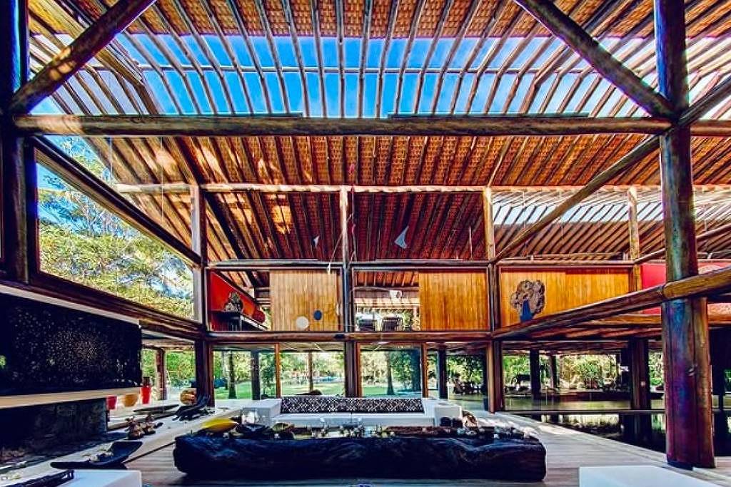 A imagem mostra o interior de uma casa moderna com estrutura aberta e grandes janelas. O teto é feito de madeira com ripas expostas, permitindo a entrada de luz natural. O espaço é amplo, com uma área de estar que inclui sofás e mesas. Ao fundo, há uma vista para um jardim ou área externa verde. As paredes são de madeira e há elementos decorativos coloridos.