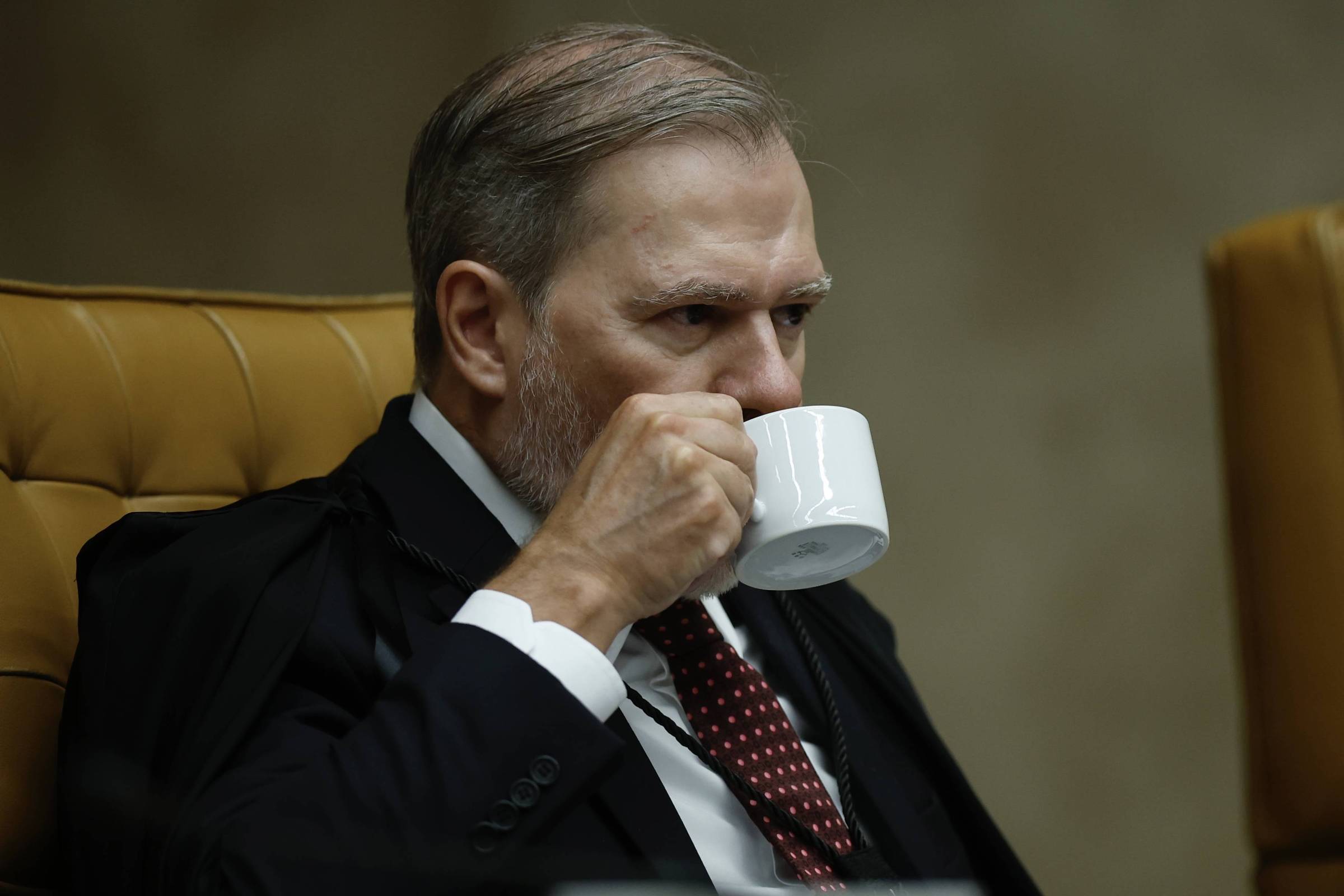 Homem de meia-idade com cabelo grisalho e barba curta, vestido com terno escuro, camisa branca e gravata vermelha com pontos brancos, sentado em cadeira de couro marrom, segurando e bebendo de uma xícara branca.
