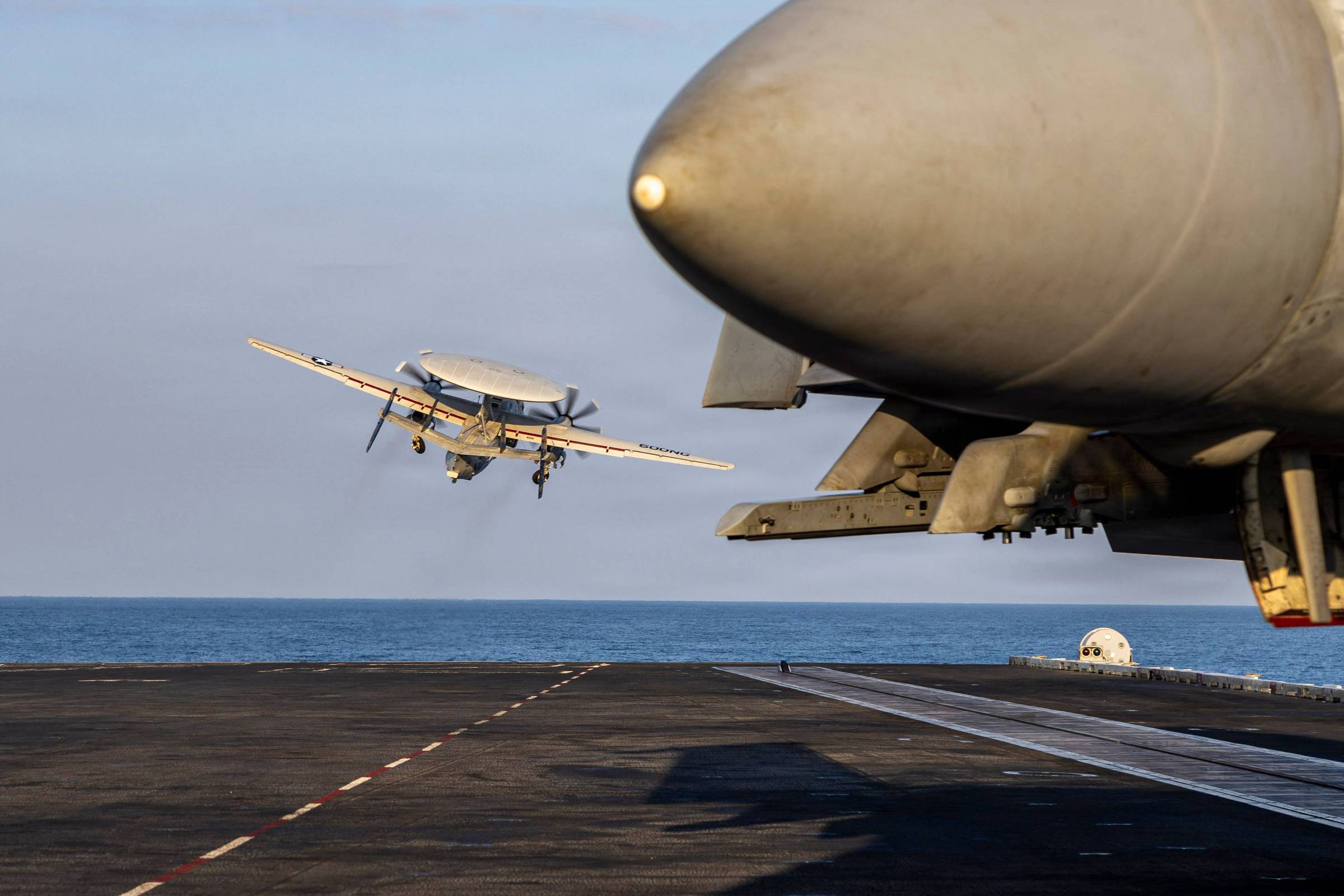 Avião decolando da pista de um porta-aviões sobre o mar, com caça estacionado em primeiro plano à direita. Céu claro e mar calmo ao fundo.