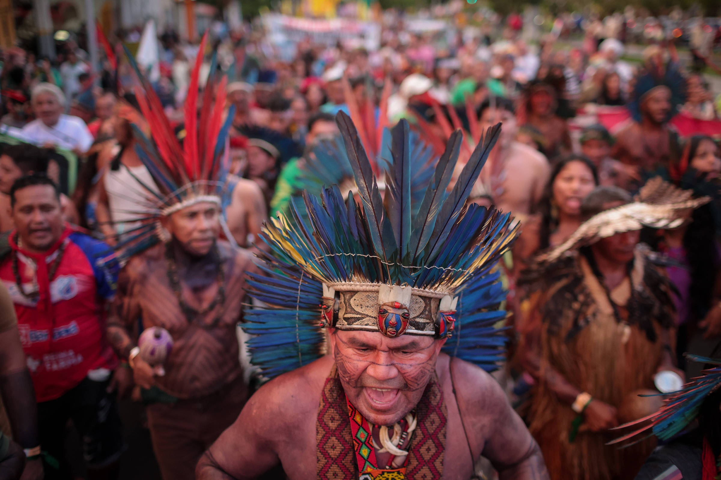 Grupo numeroso de indígenas participa de manifestação ao ar livre, muitos usando cocares de penas coloridas e trajes tradicionais. Homem em primeiro plano usa cocar azul e vermelho e expressa intensidade vocal. Pessoas ao fundo também vestem roupas típicas e seguram instrumentos ou objetos.