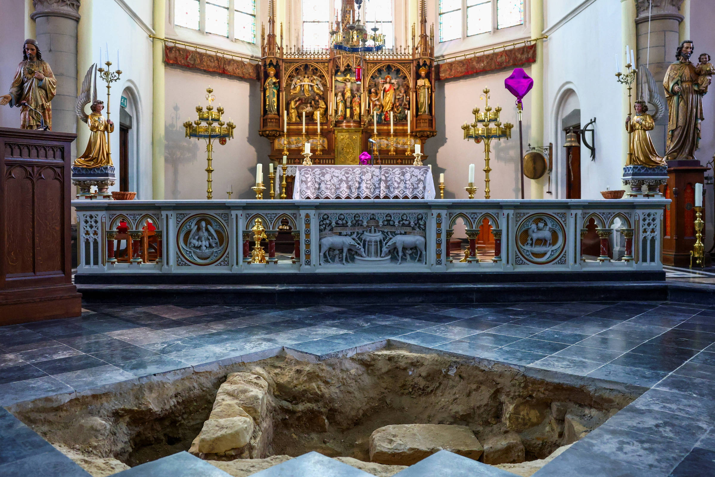 Altar decorado com esculturas douradas e castiçais em igreja iluminada por janelas altas. No chão de pedra à frente, há uma escavação arqueológica retangular com restos de estruturas antigas visíveis.