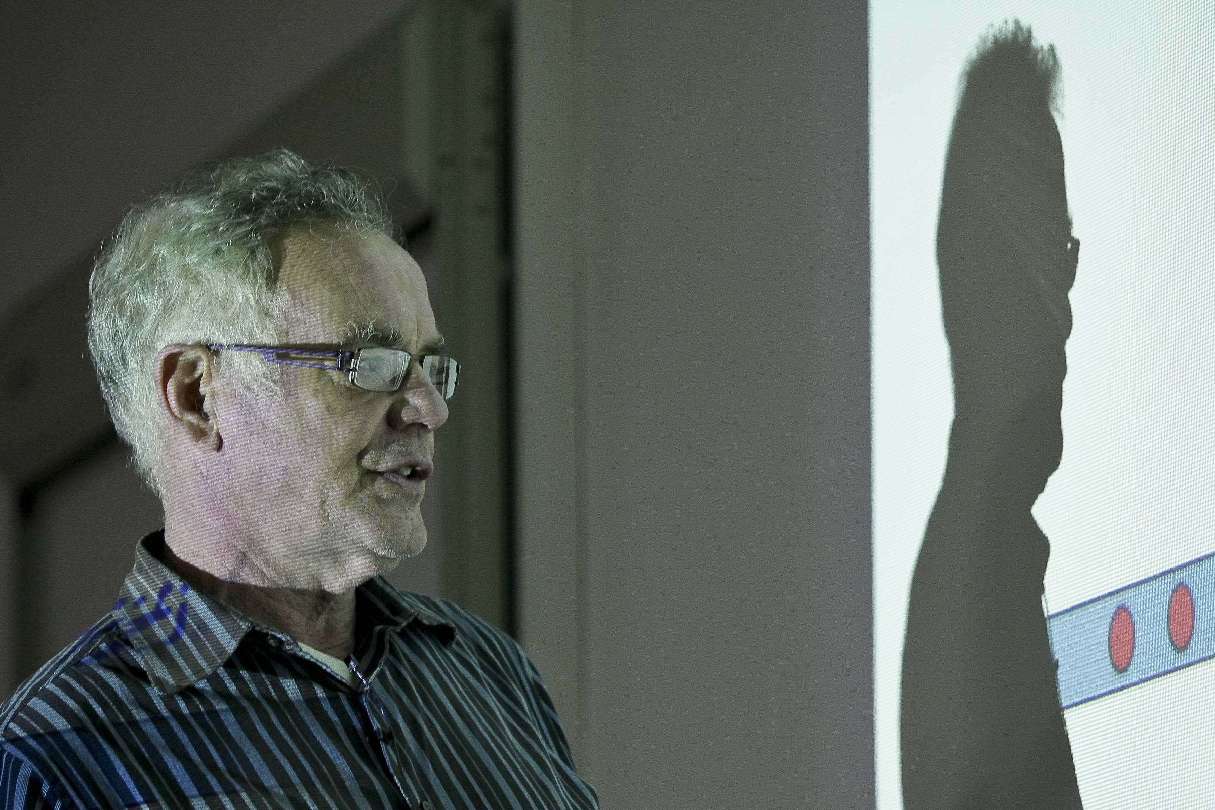 Homem idoso de cabelos grisalhos e óculos fala enquanto está de perfil, com camisa listrada, diante de uma parede onde sua sombra é projetada. A sombra mostra detalhes do rosto e dos óculos, e parte de uma imagem com círculos vermelhos em faixa azul aparece na projeção.