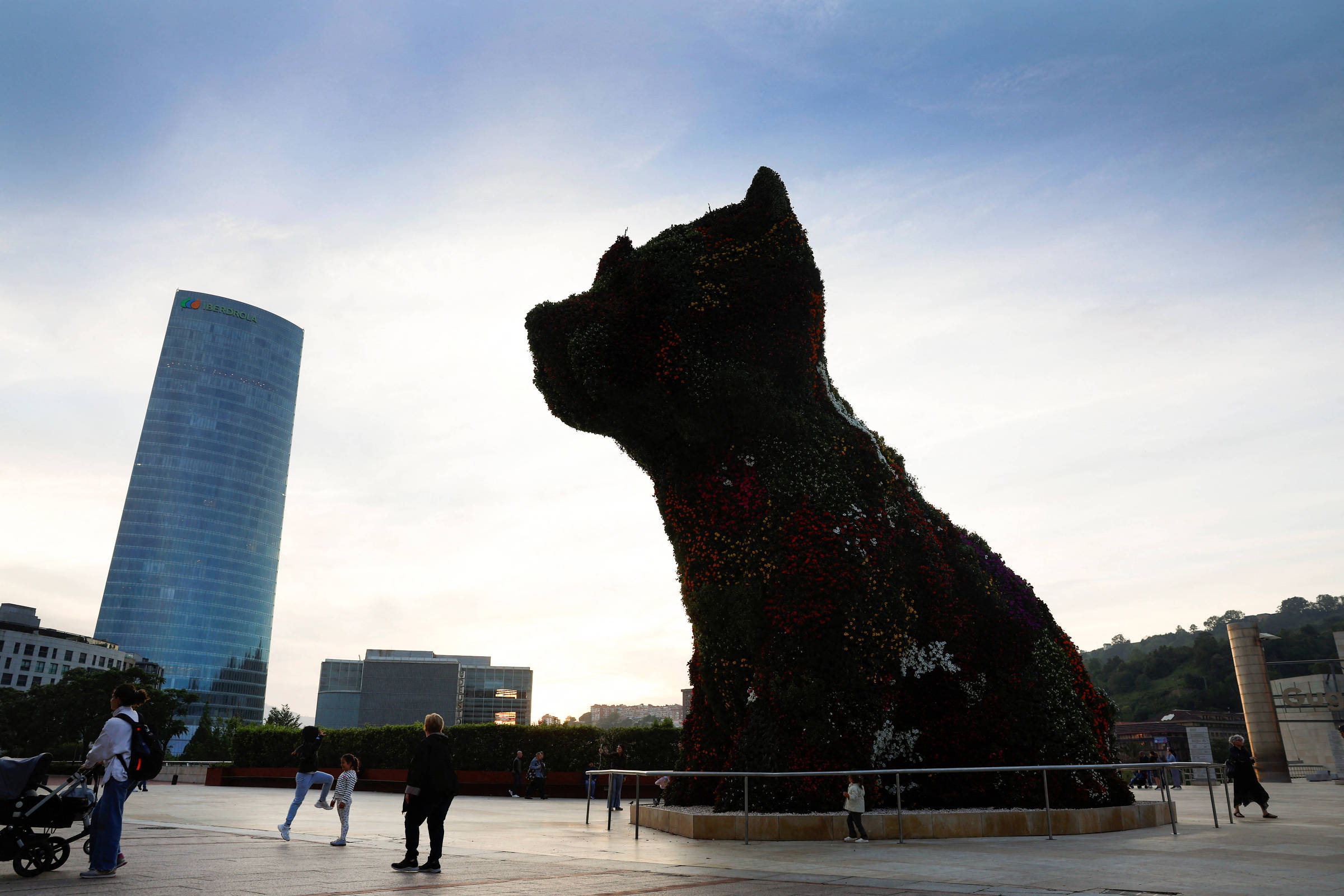 Escultura monumental em forma de cachorro feita com plantas e flores, localizada em praça ampla com pessoas ao redor. Ao fundo, edifícios modernos e céu claro ao entardecer.