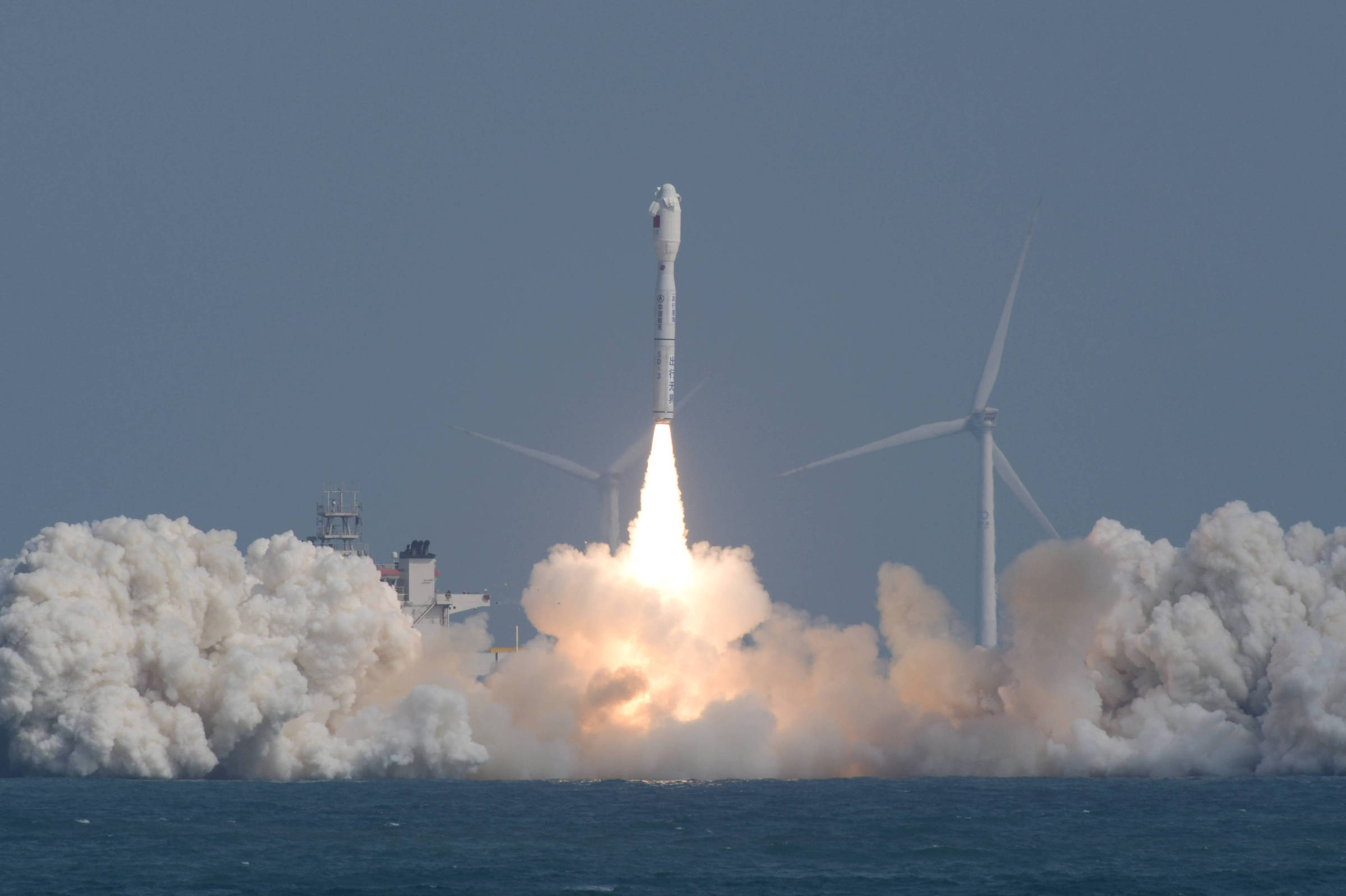 Foguete decolando da plataforma próxima ao mar, com chamas intensas e grande nuvem de fumaça branca ao redor. Ao fundo, duas turbinas eólicas visíveis sob céu claro.