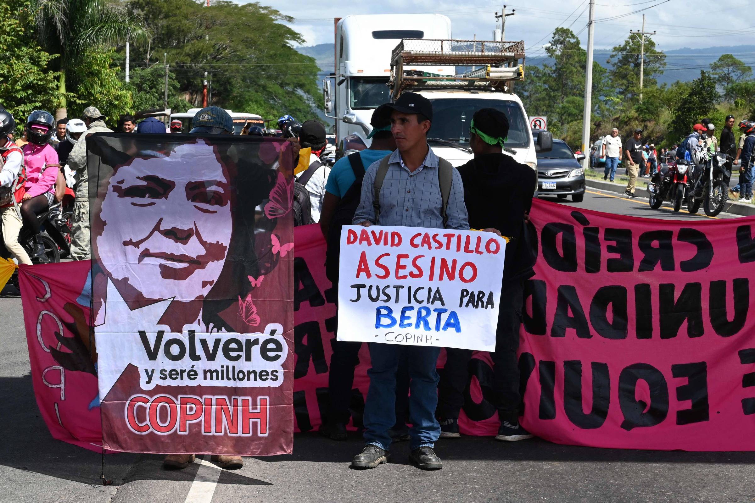 Manifestantes bloqueiam estrada segurando faixas e cartazes. Um homem no centro segura placa com texto