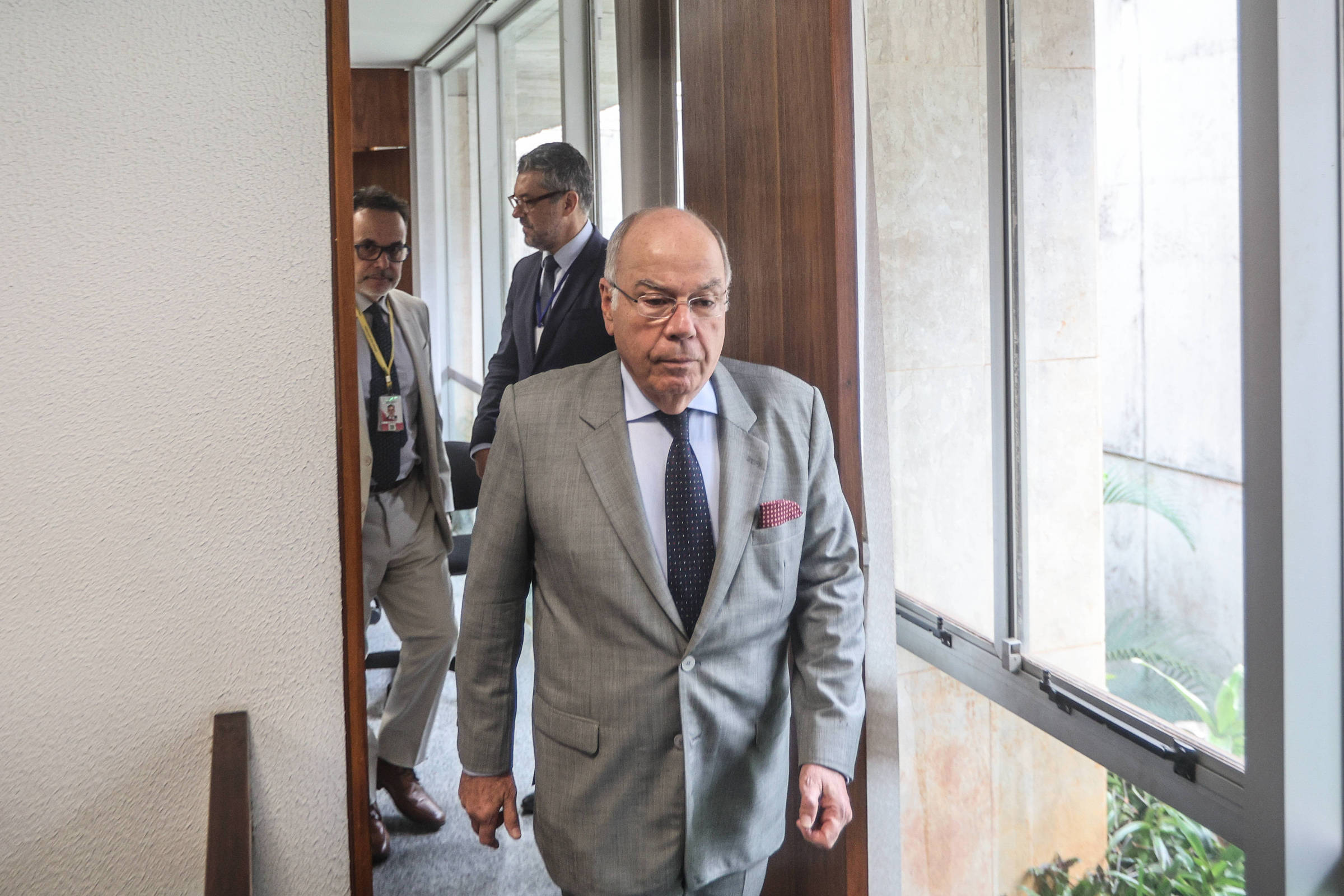 Homem de terno cinza claro, camisa azul e gravata escura caminha por corredor interno com janelas à direita. Dois homens ao fundo, um com crachá, observam a passagem.
