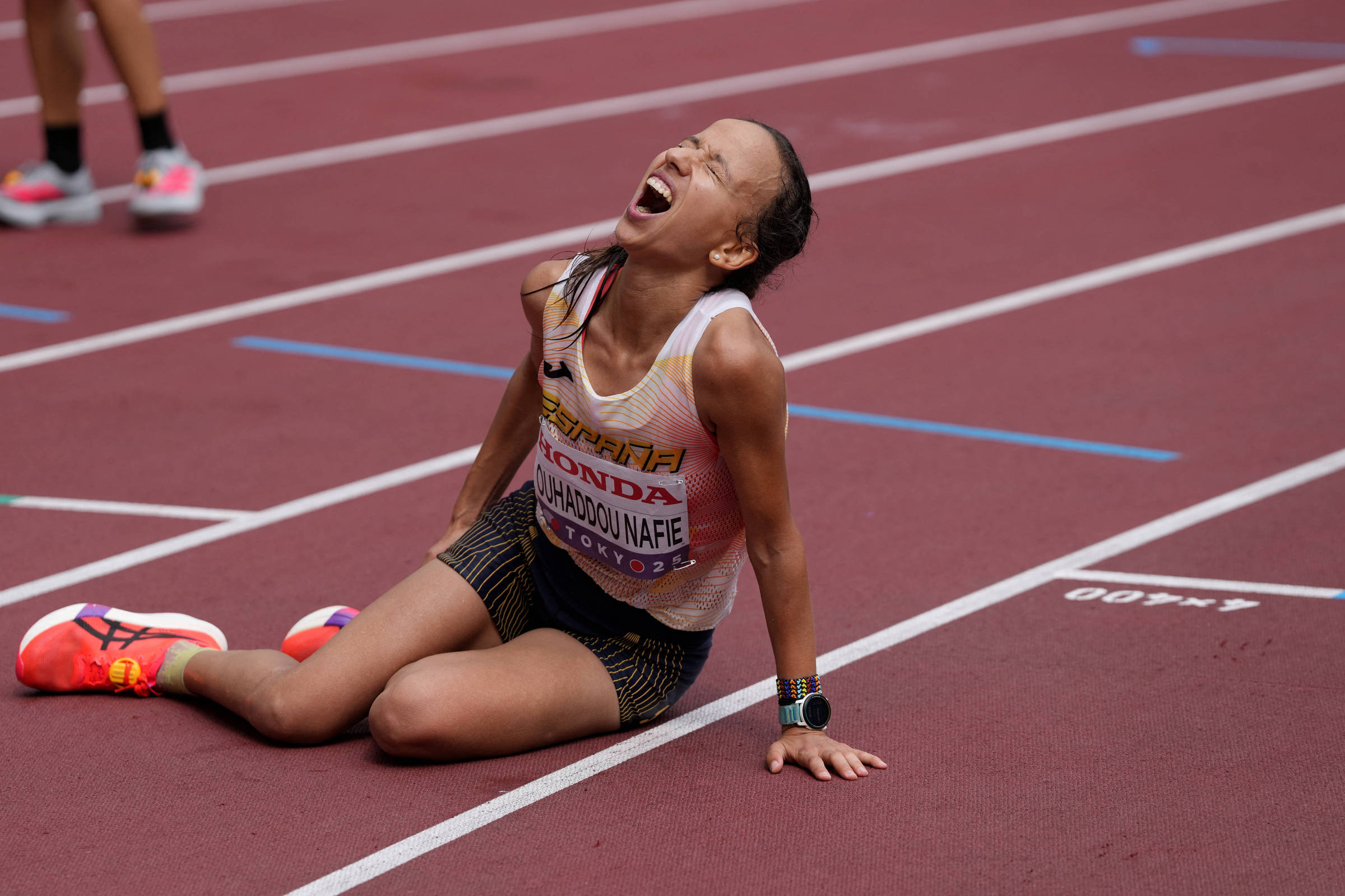 Atleta feminina sentada no chão da pista de atletismo, com expressão de esforço ou dor, vestindo uniforme branco e preto com o nome