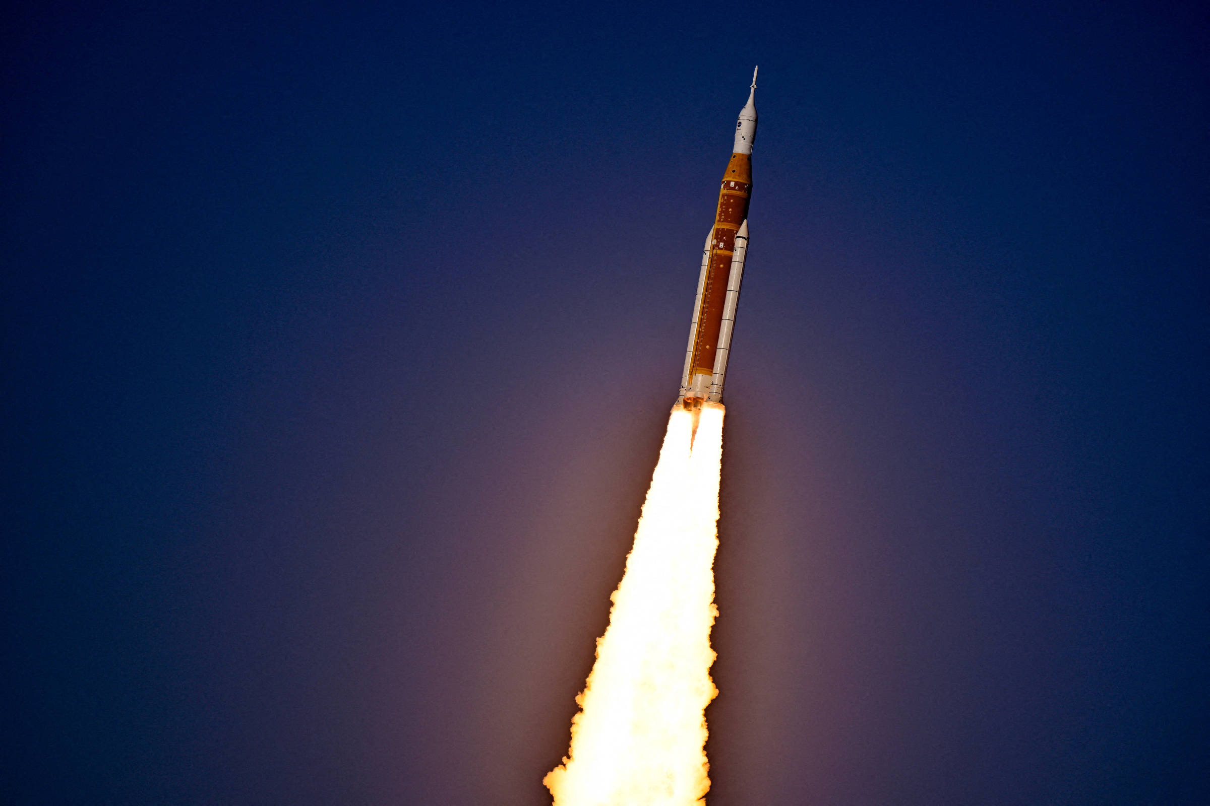 Foguete em ascensão com chamas intensas saindo da base, contra céu azul escuro. Trajeto vertical indica decolagem recente.