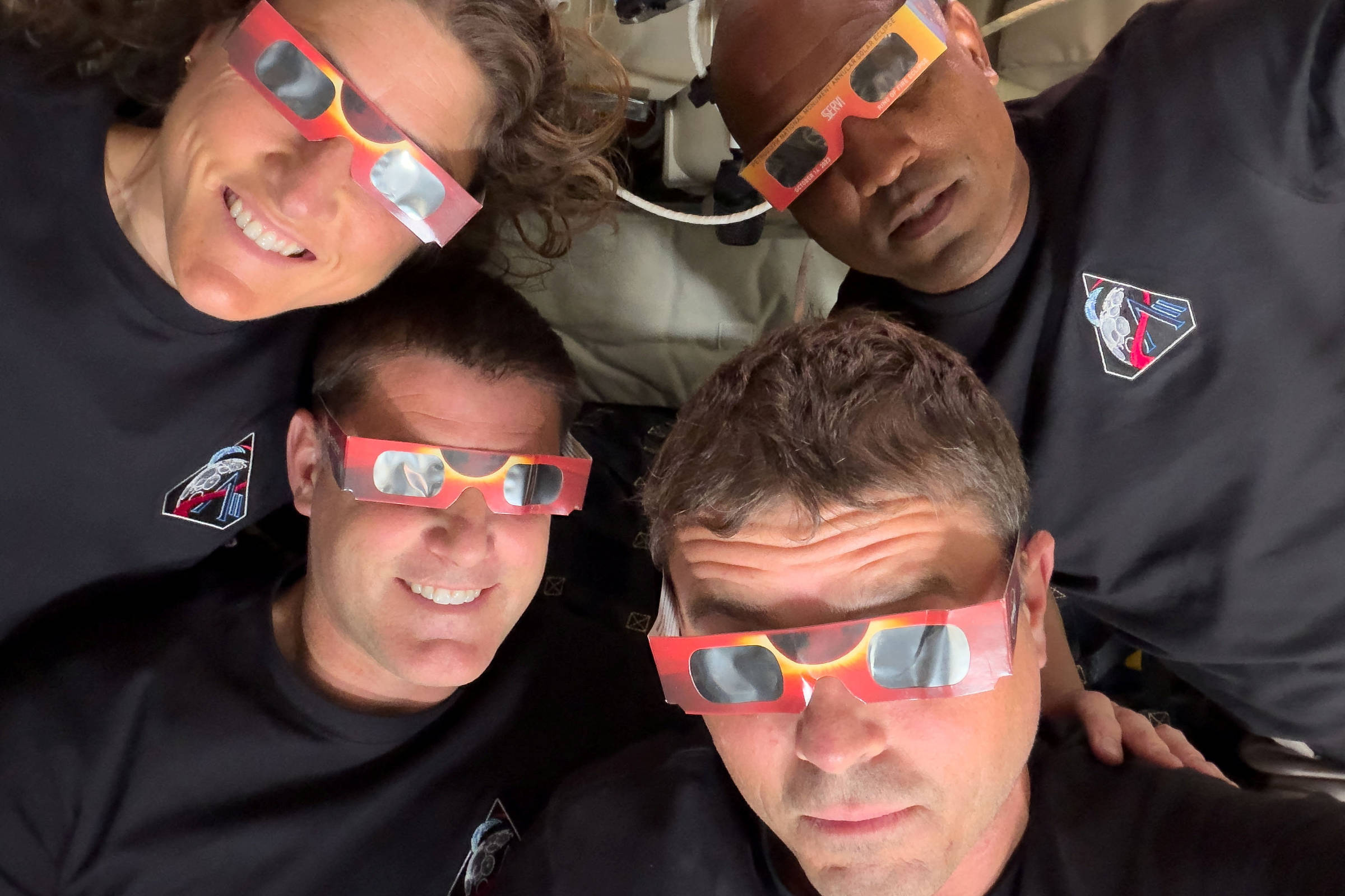 Quatro astronautas deitados lado a lado, usando óculos vermelhos para observação de eclipse. Todos vestem camisetas pretas com emblema da missão espacial.
