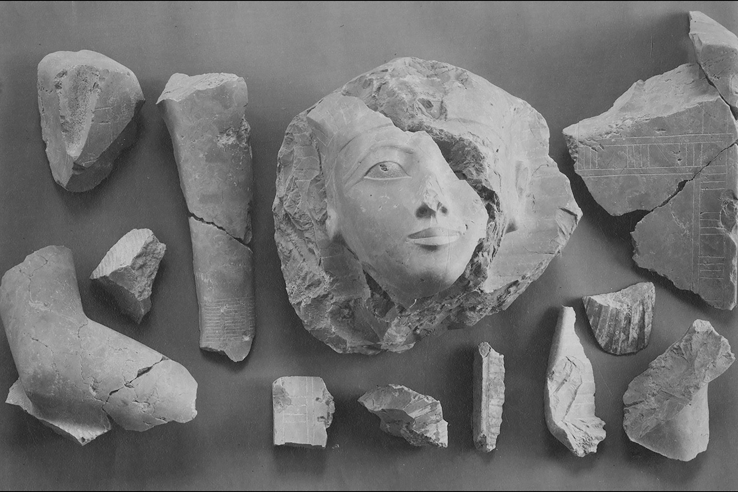 Fragmentos de pedra dispostos sobre superfície plana, destacando um rosto feminino esculpido parcialmente preservado no centro, com olhos, nariz e boca visíveis. Ao redor, pedaços irregulares de pedra com marcas e entalhes, sugerindo partes de uma escultura maior.