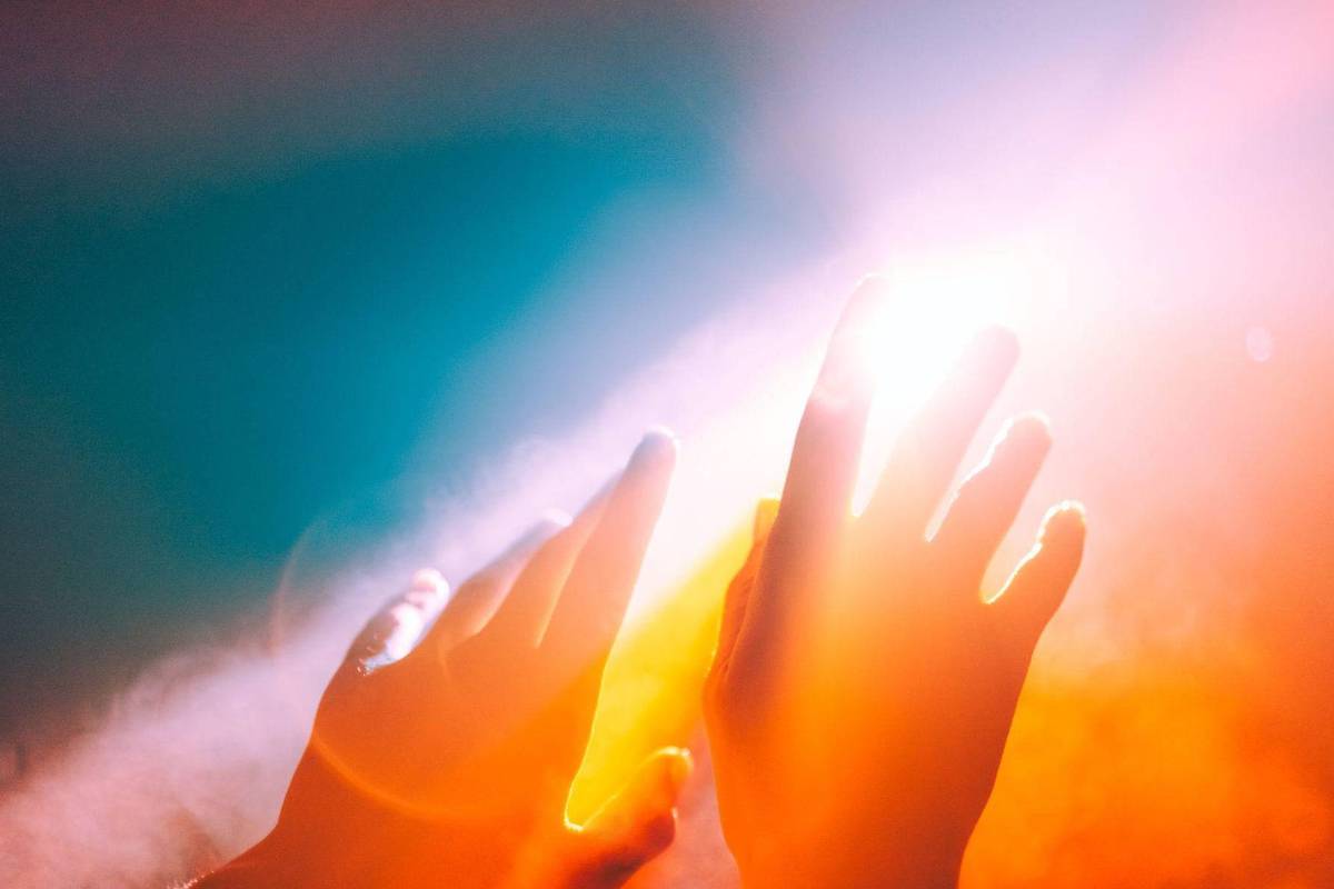 Duas mãos abertas e estendidas em direção a uma fonte de luz intensa e difusa, com tons quentes de laranja e amarelo, contrastando com um fundo azul esmaecido.