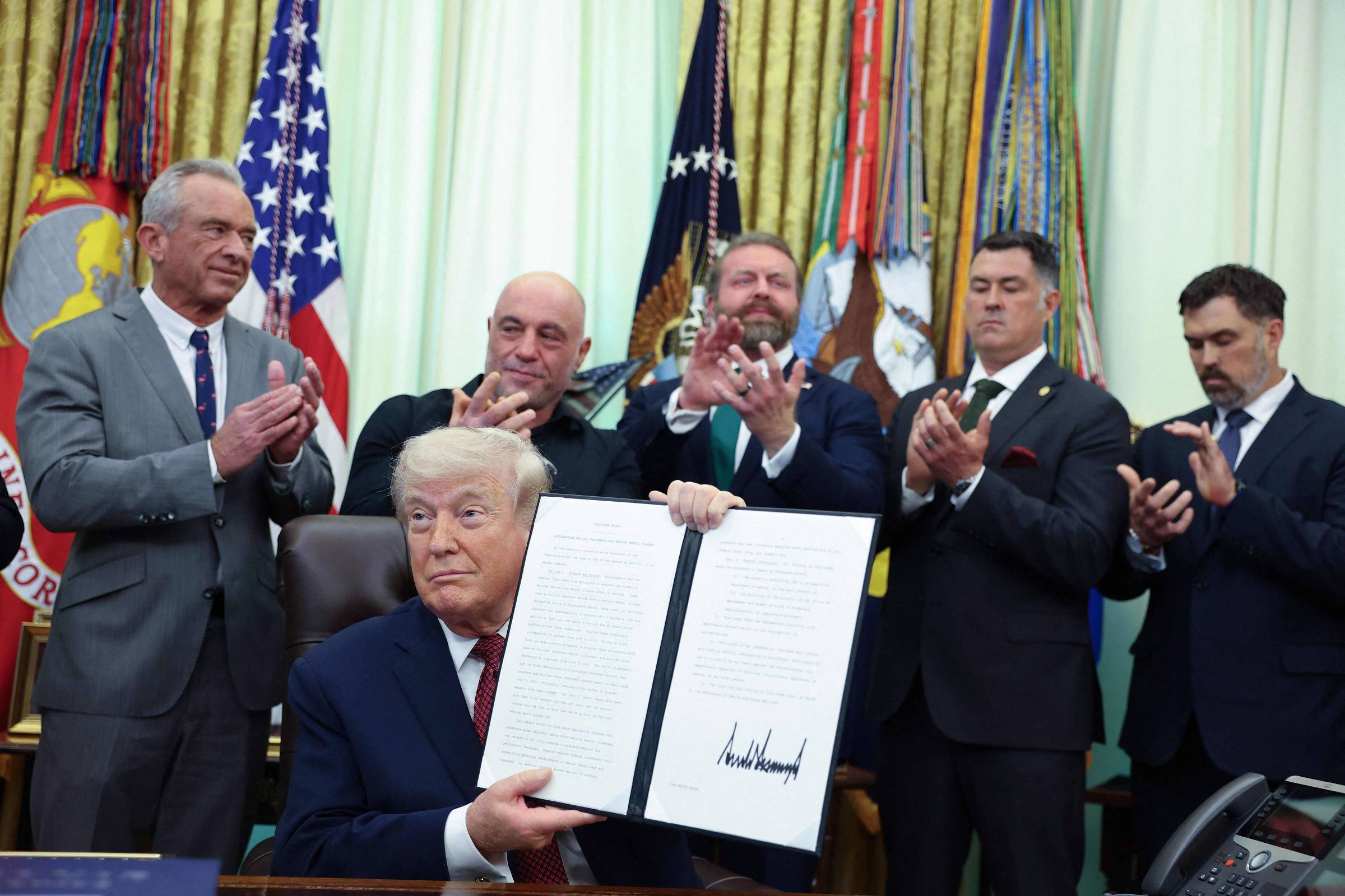 Ex-presidente dos Estados Unidos sentado na mesa do Salão Oval, segurando documento assinado. Cinco homens em pé ao fundo aplaudem, com bandeiras americanas e militares atrás.