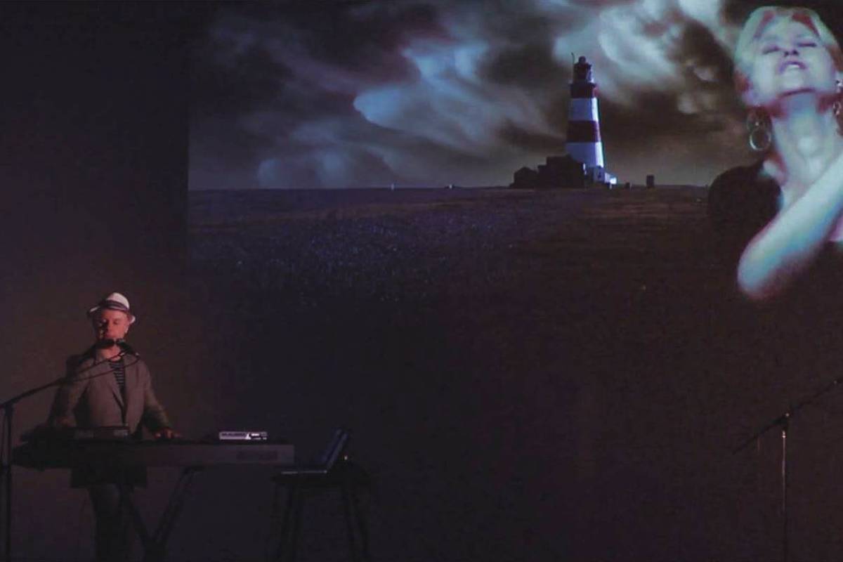 Músico toca teclado à esquerda em ambiente escuro. Ao fundo, projeção mostra farol em paisagem noturna e rosto de mulher cantando com olhos fechados.
