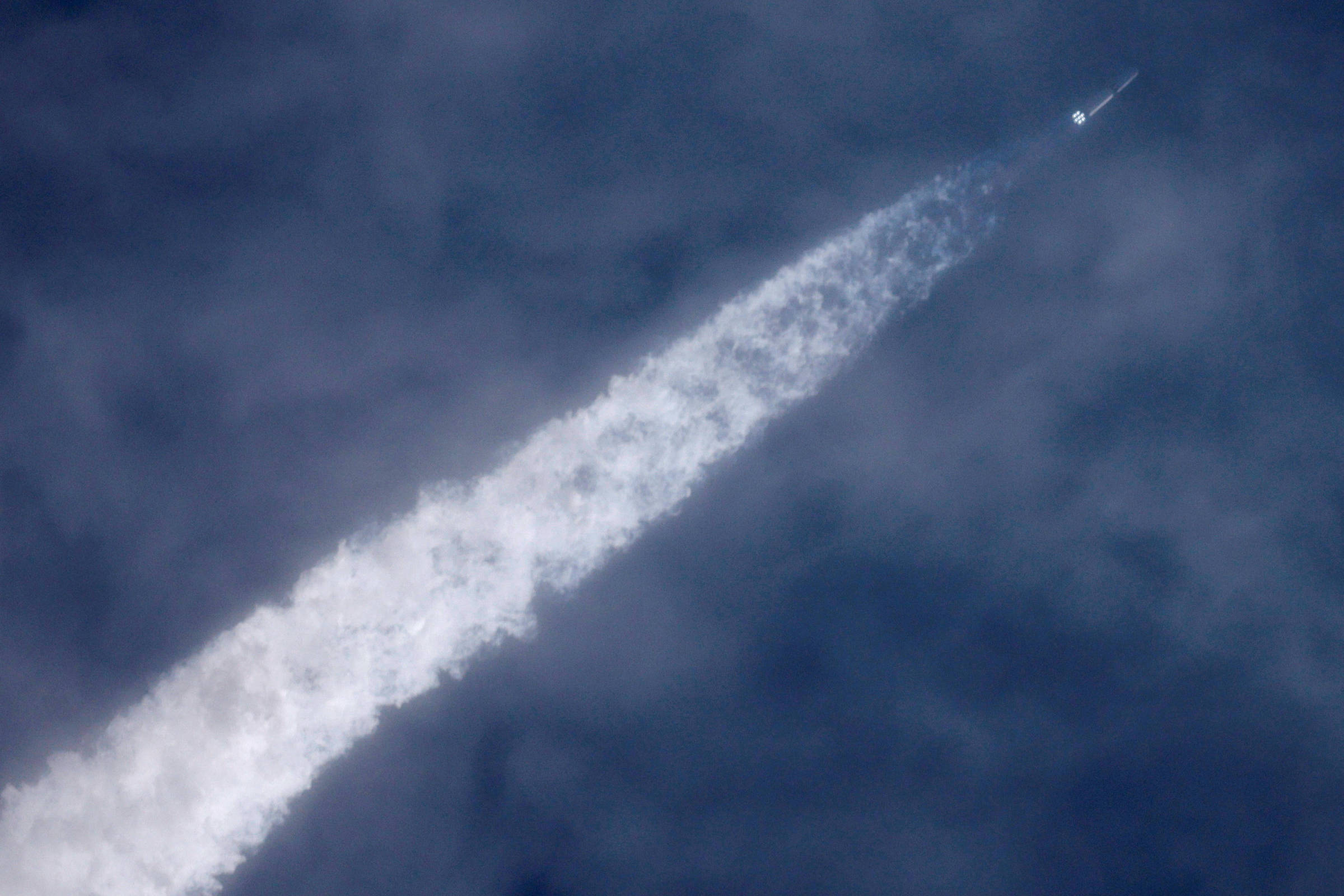Foguete branco sobe em direção ao céu azul com nuvens, deixando uma longa trilha espessa de fumaça branca.