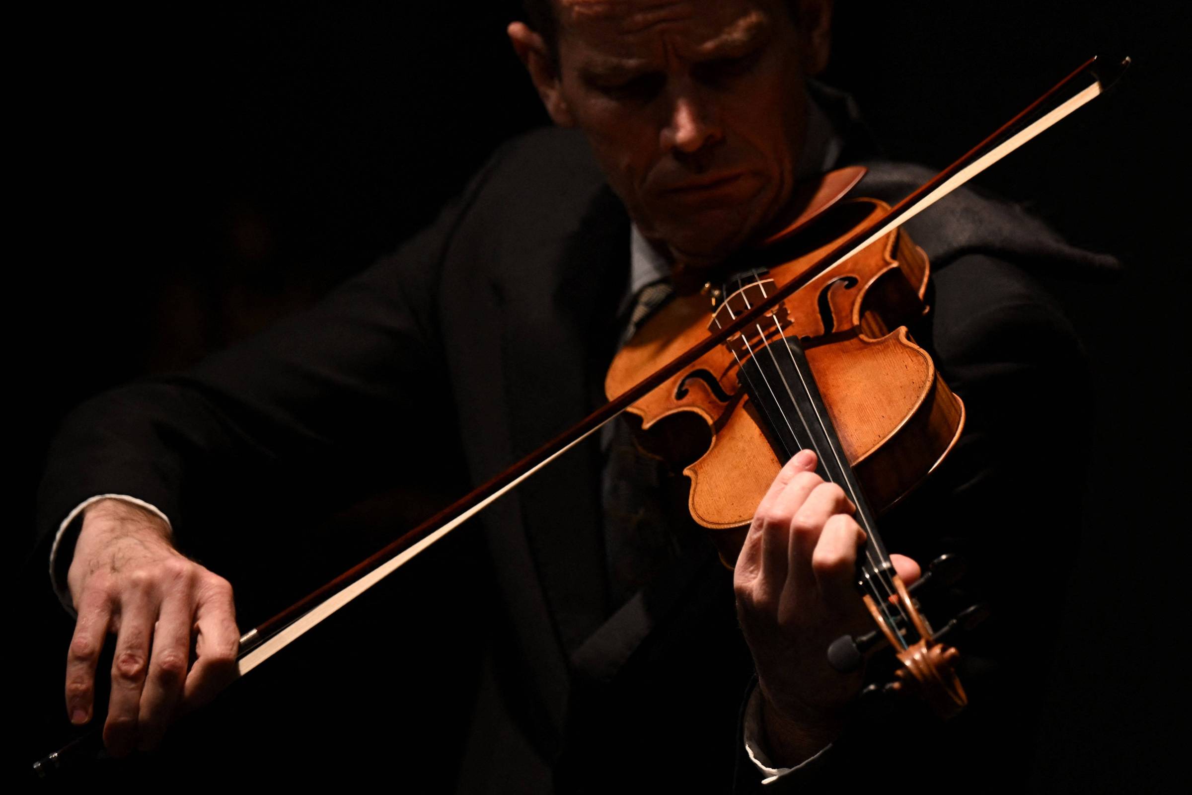 Homem vestido com terno escuro toca violino com expressão concentrada. Fundo preto destaca o instrumento e as mãos do músico.