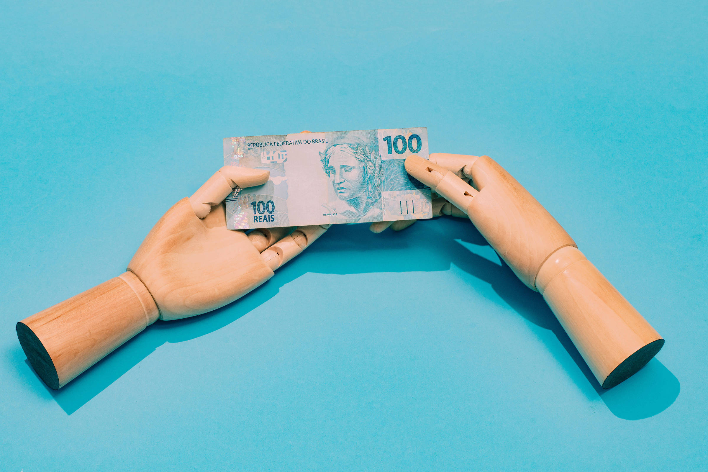 A imagem mostra duas mãos de madeira, uma de cada lado, segurando uma nota de 100 reais. O fundo é azul claro, destacando a nota que possui a figura de uma mulher e o valor impresso em destaque. As mãos estão posicionadas de forma a parecer que estão trocando a nota.