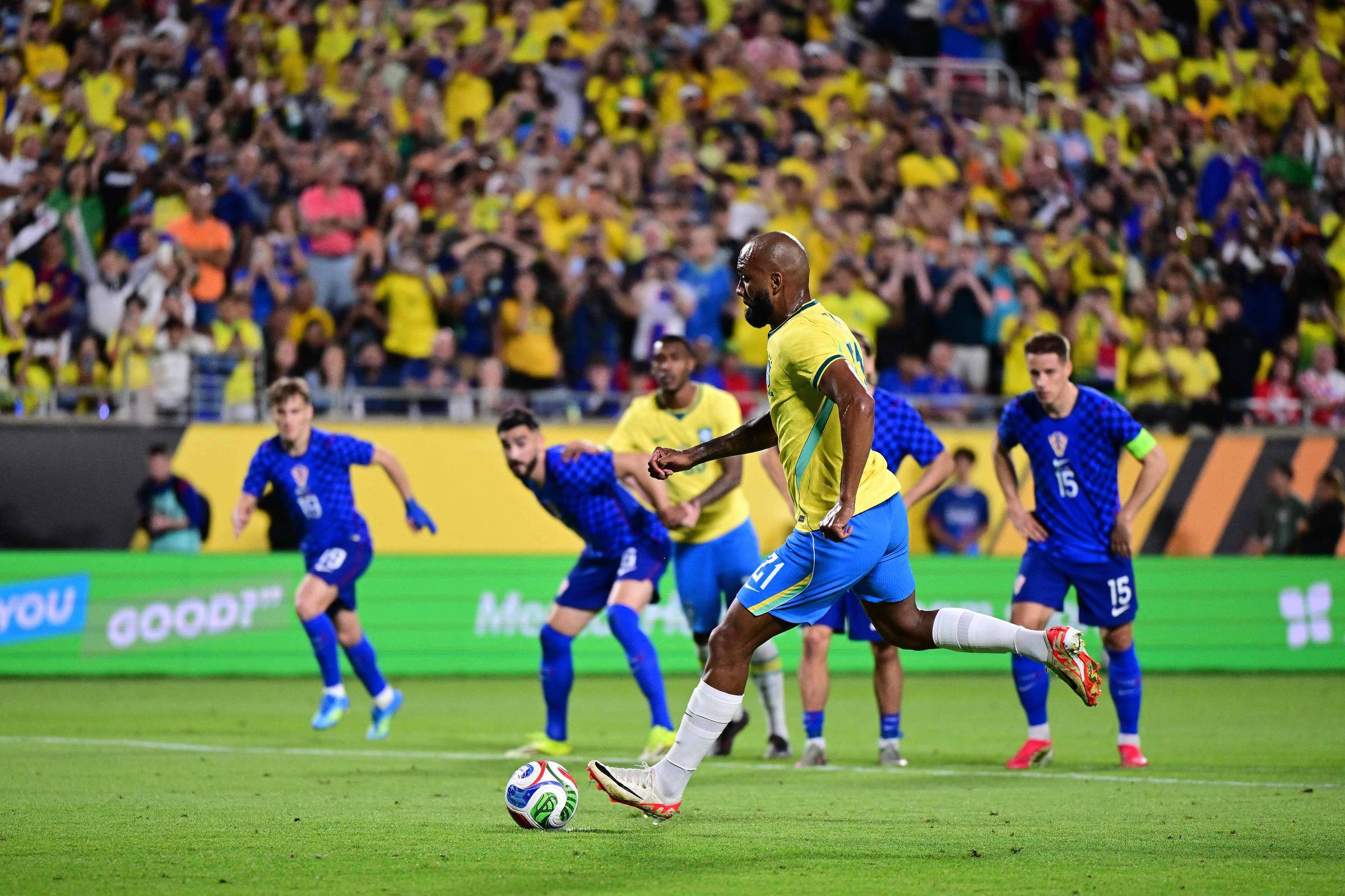 Jogador brasileiro com camisa amarela e calção azul executa cobrança de pênalti em campo gramado. Quatro jogadores adversários de azul aguardam atrás da linha da bola, enquanto torcida lota arquibancada ao fundo.