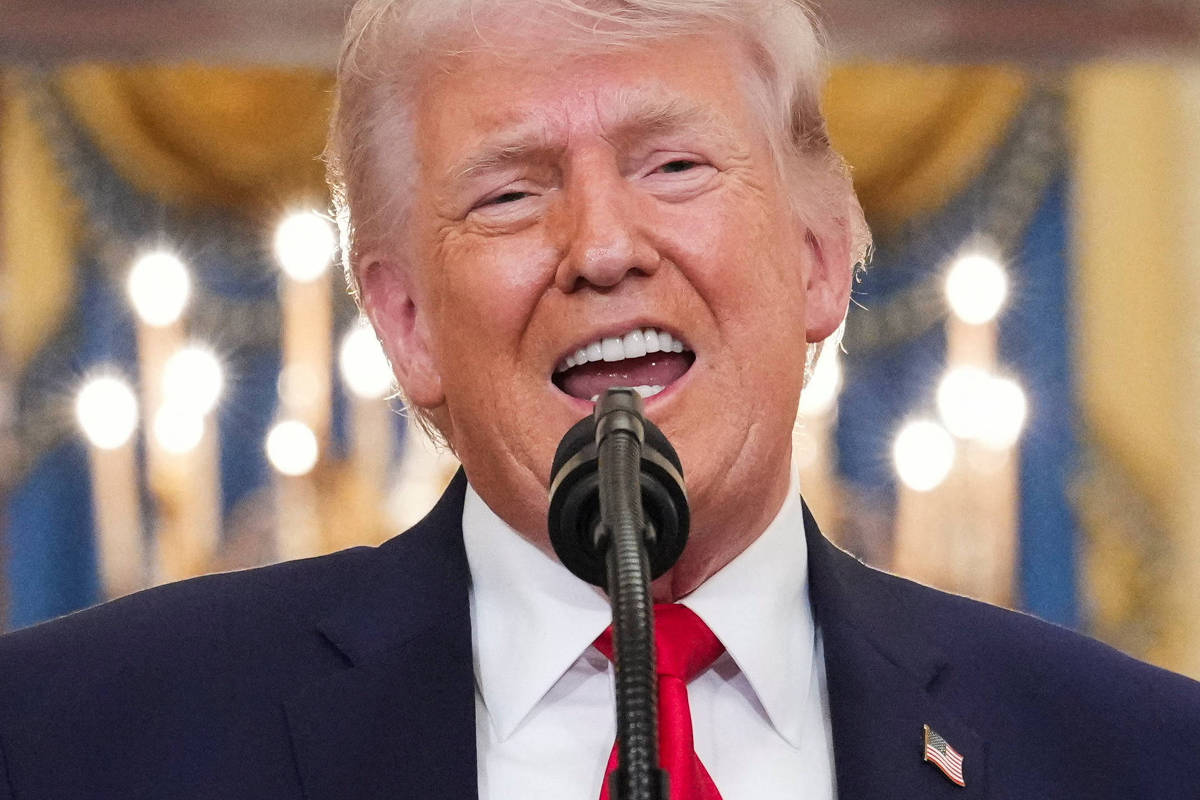 Homem branco de cabelos grisalhos fala ao microfone em ambiente interno com iluminação e cortinas douradas ao fundo. Ele veste terno escuro, camisa branca e gravata vermelha, com broche da bandeira dos EUA na lapela.