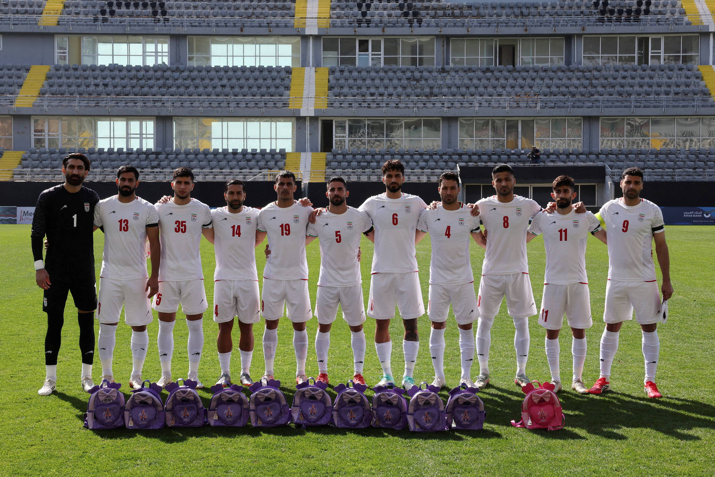 Onze jogadores de futebol alinhados no gramado de um estádio vazio, vestindo uniformes brancos com números vermelhos. Na frente deles, dez mochilas roxas e uma rosa estão dispostas no chão.