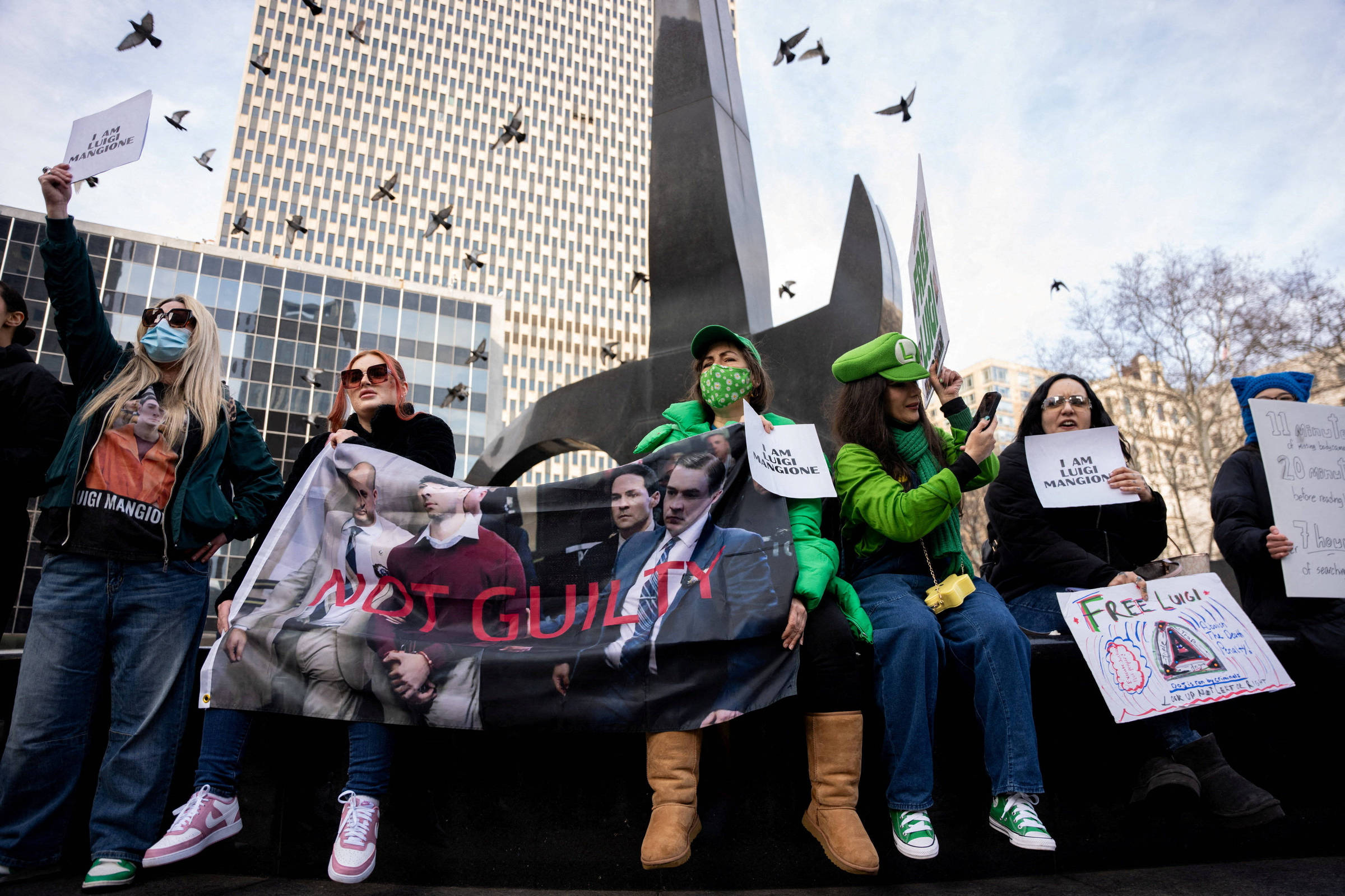 Grupo de seis manifestantes sentados em escadaria urbana diante de prédio alto. Quatro seguram cartazes, incluindo um com foto de três homens e texto