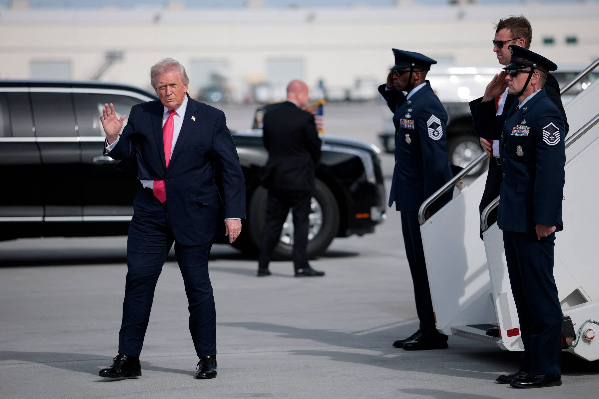 Ex-presidente dos EUA caminha na pista de aeroporto com terno escuro e gravata vermelha, acenando com a mão direita. Dois militares em uniforme azul escuro fazem continência próximos à escada de aeronave, enquanto outro homem de terno observa ao fundo.