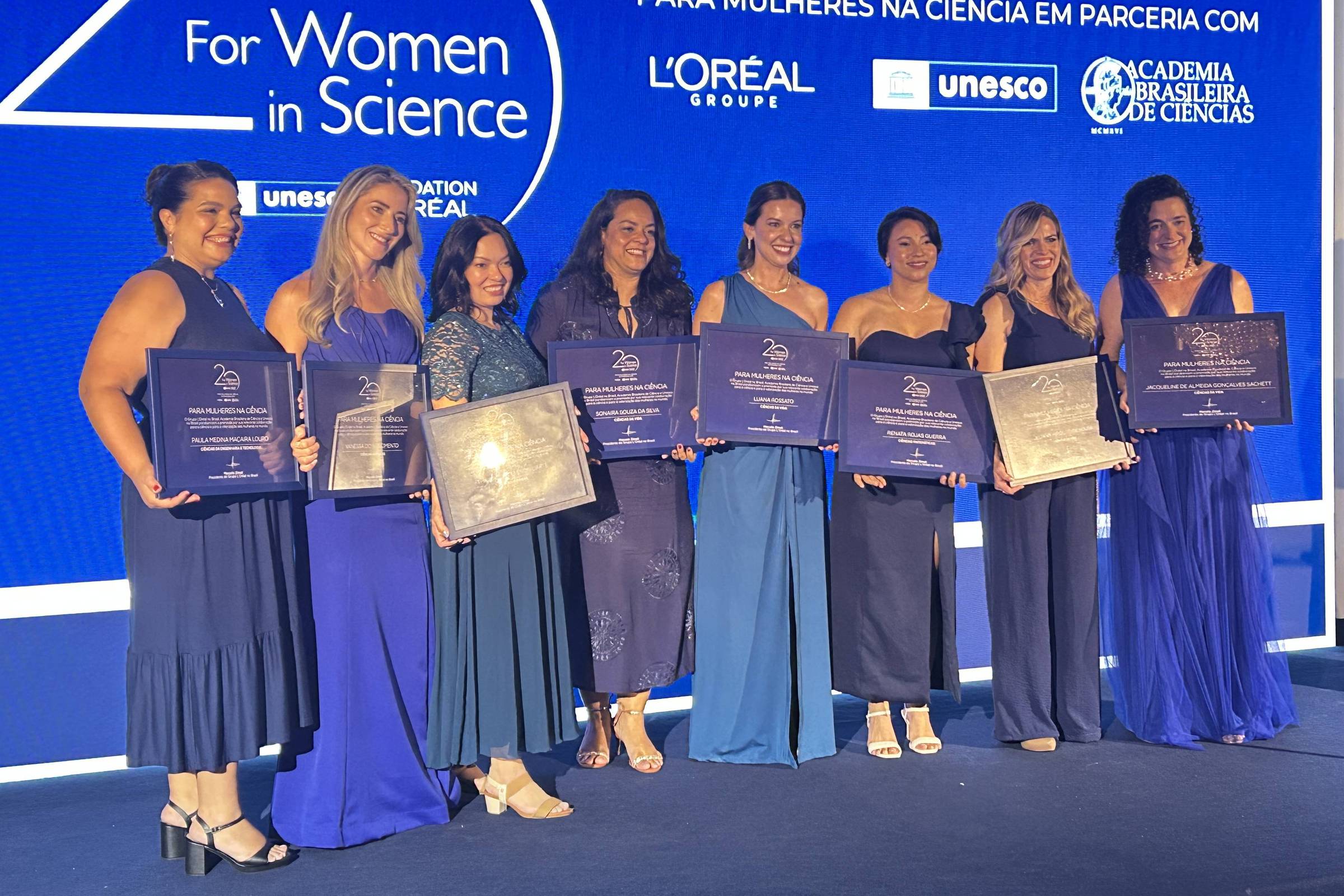 Sete mulheres vestidas com roupas formais em tons de azul posam em palco segurando placas de premiação. Ao fundo, painel azul com logotipos da L