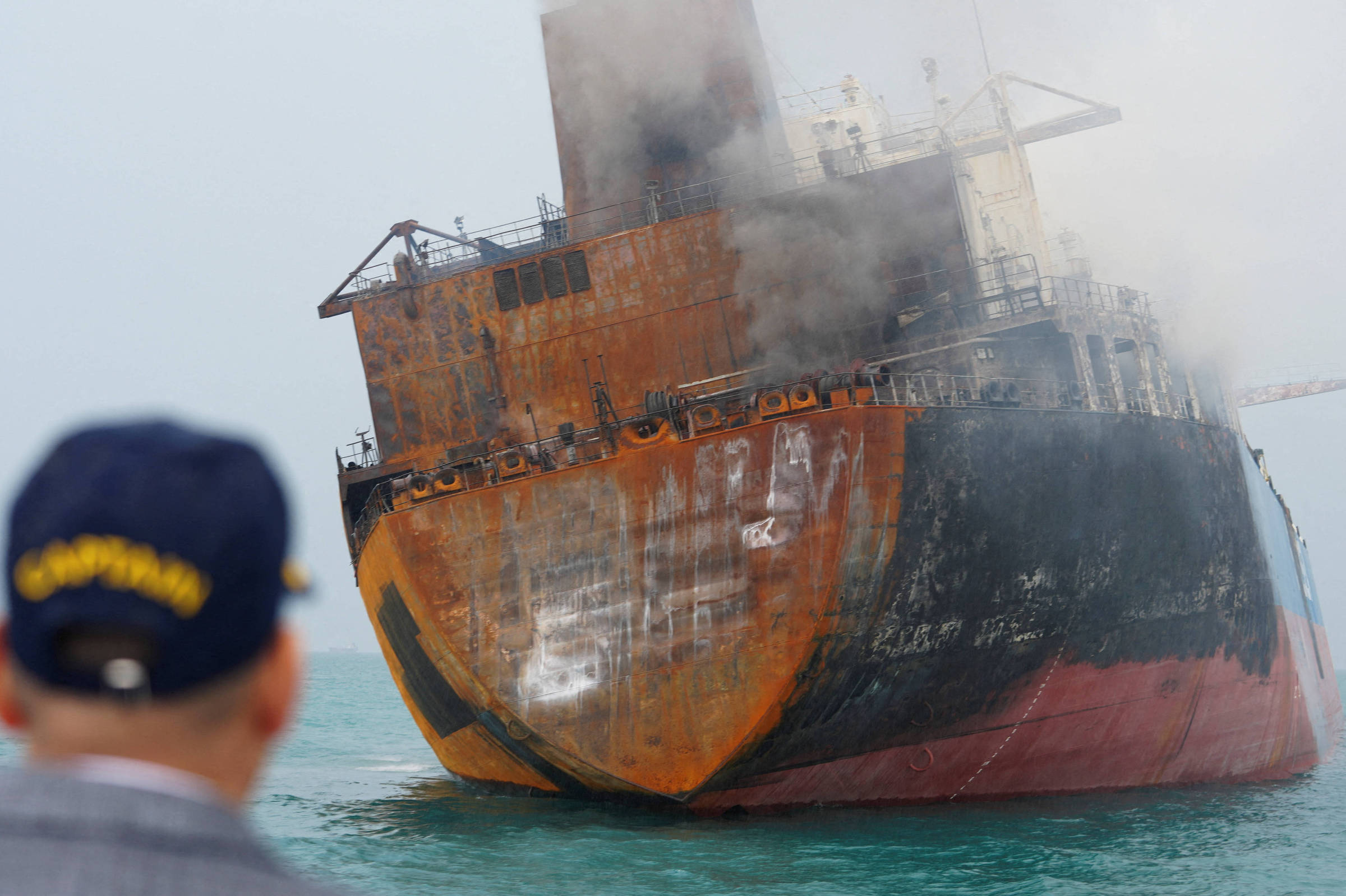 Navio grande com casco enferrujado e fumaça saindo da parte superior flutua no mar. Homem de costas usa boné azul com amarelo e observa a embarcação.