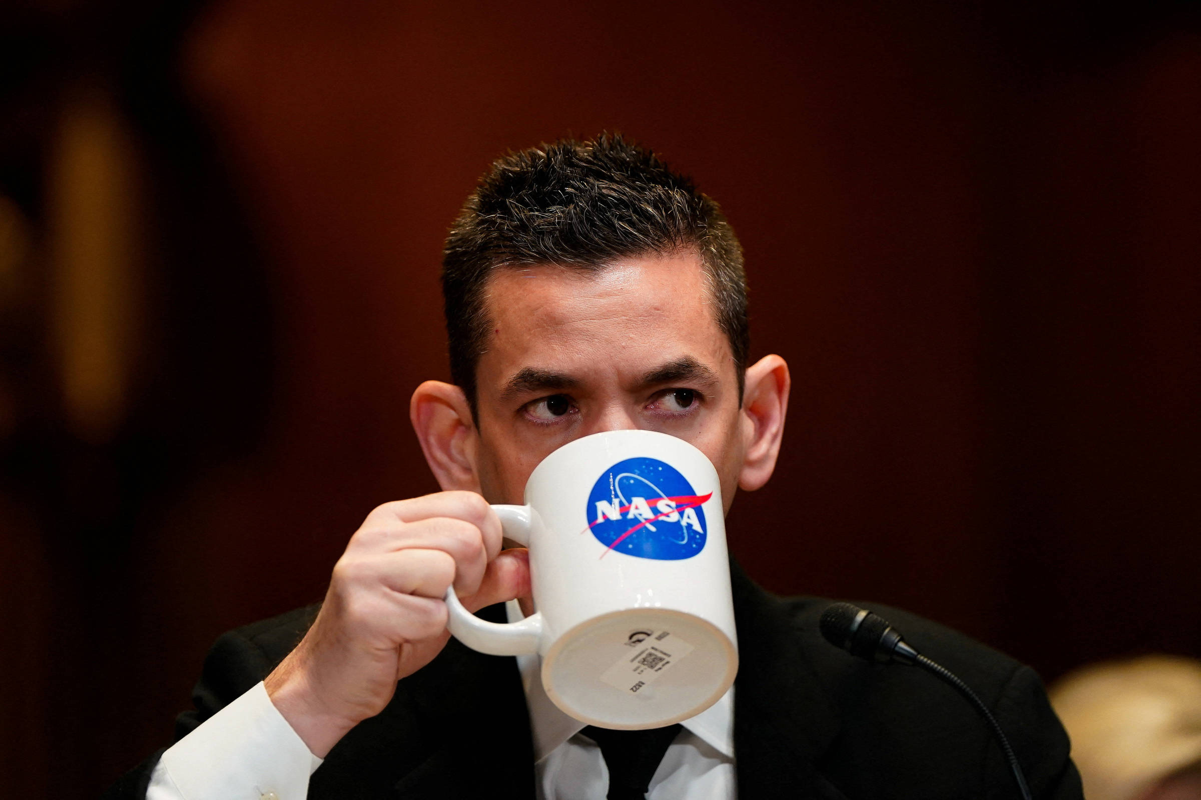 Homem de terno preto e camisa branca segura e bebe de uma caneca branca com o logo da NASA em azul e vermelho. Ele está em ambiente interno com fundo desfocado e microfone à sua frente.