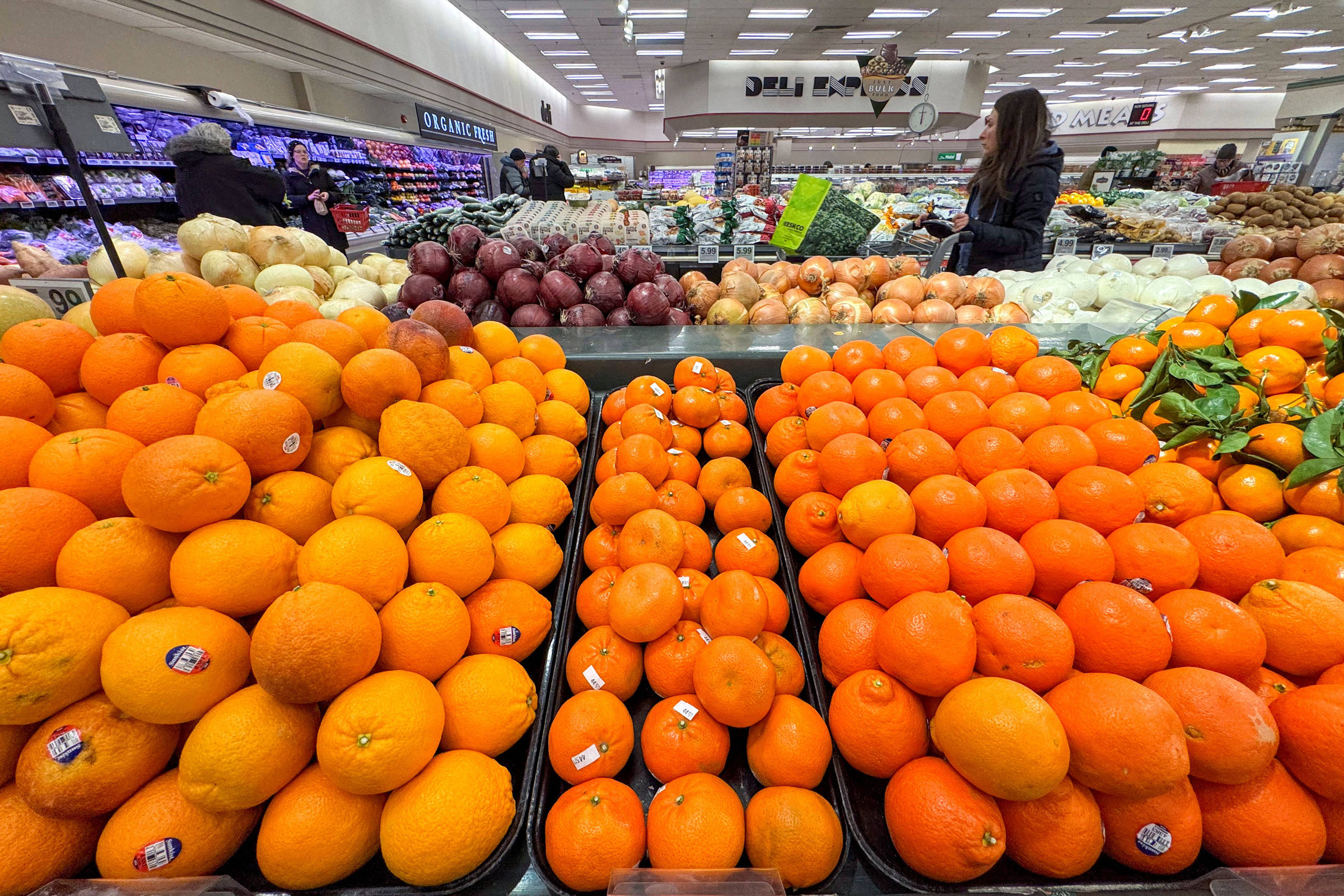 Bancada de supermercado com laranjas, tangerinas e outros cítricos em primeiro plano. Ao fundo, cebolas roxas, cebolas brancas e outros vegetais empilhados. Pessoas circulam entre as gôndolas em ambiente iluminado.
