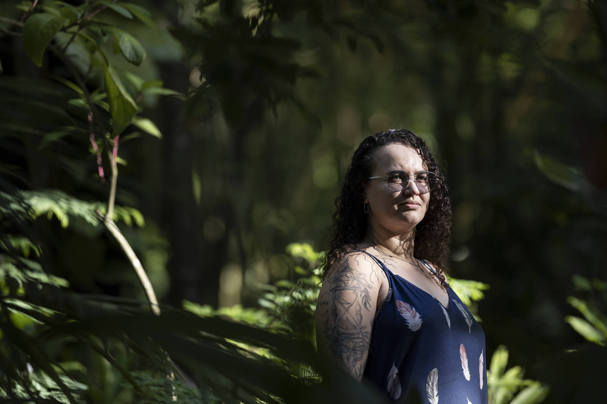 Mulher de cabelos cacheados e óculos está em pé em meio a vegetação densa, com luz natural iluminando seu rosto e parte do corpo. Ela veste vestido azul com estampa de penas e tem tatuagens visíveis no braço esquerdo. Fundo desfocado mostra árvores e folhagens.
