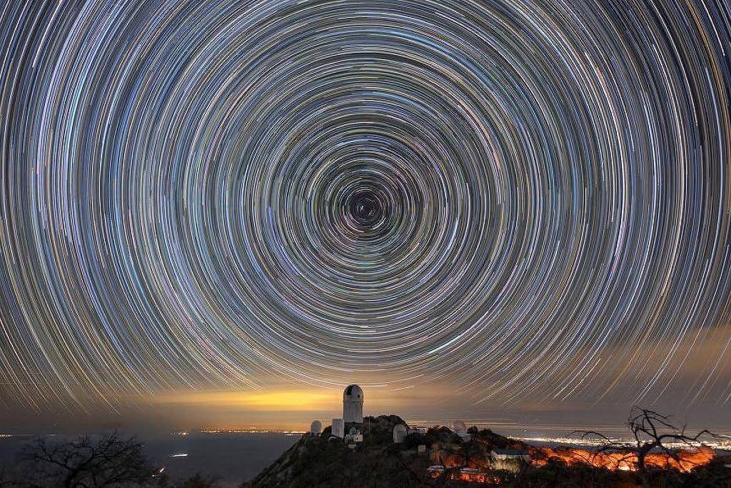 Rastros circulares de estrelas no céu noturno formam um padrão concêntrico acima de um observatório em colina. Luzes da cidade iluminam o horizonte ao fundo.