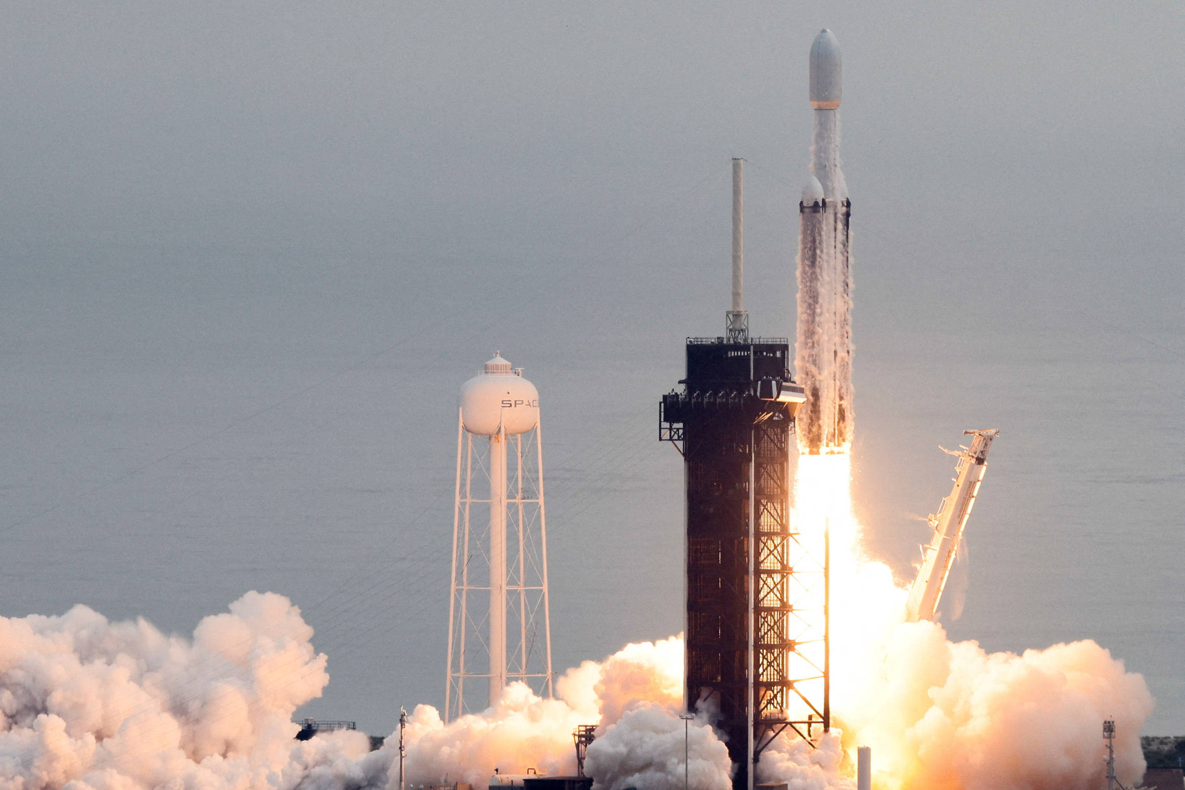 Foguete Falcon 9 decola da plataforma de lançamento com chamas intensas e fumaça densa ao redor. Torre de lançamento e reservatório de água visíveis ao fundo em ambiente claro.