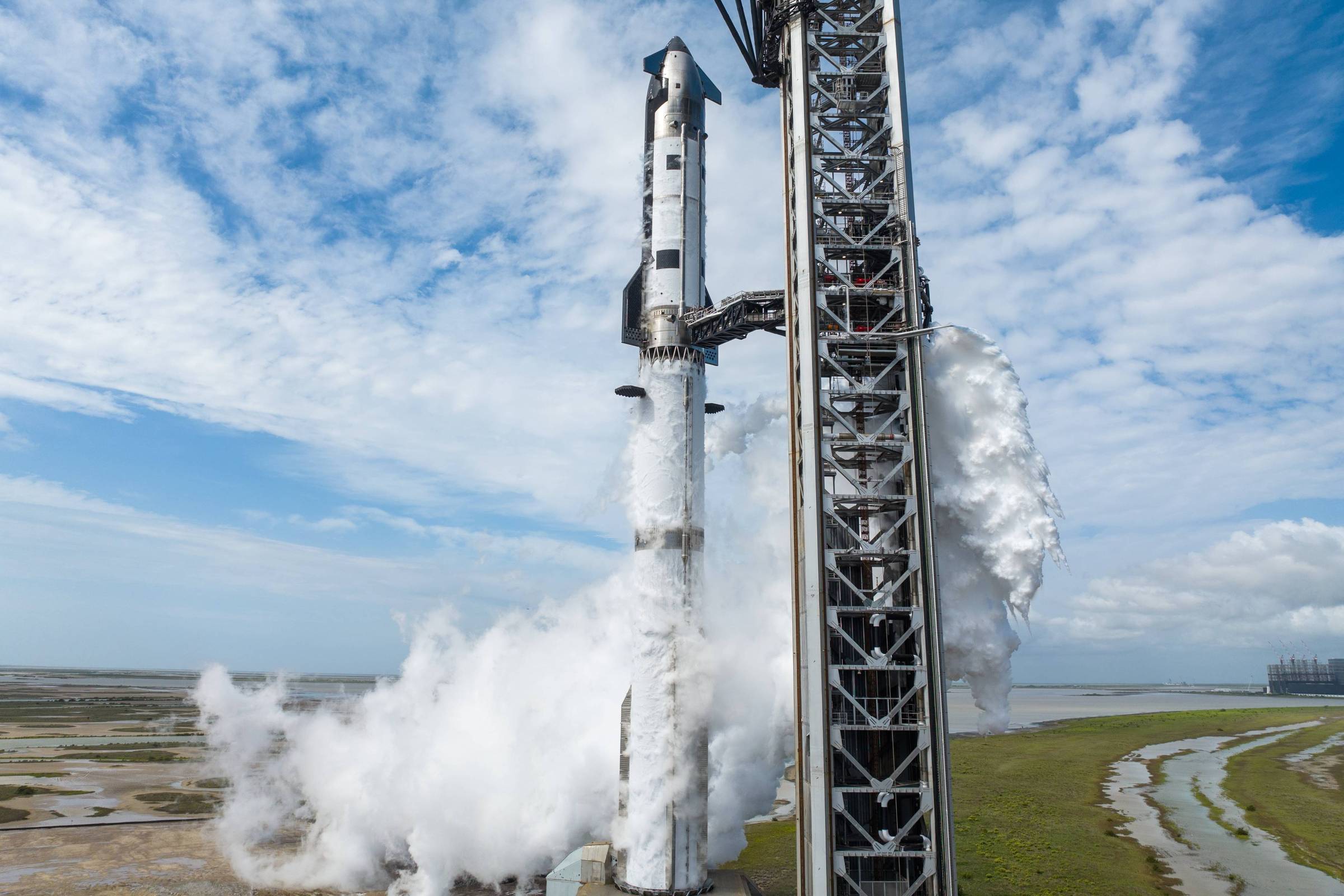 Foguete branco vertical na plataforma de lançamento com estrutura metálica ao lado. Vapor branco sai da base do foguete em ambiente aberto com céu parcialmente nublado.