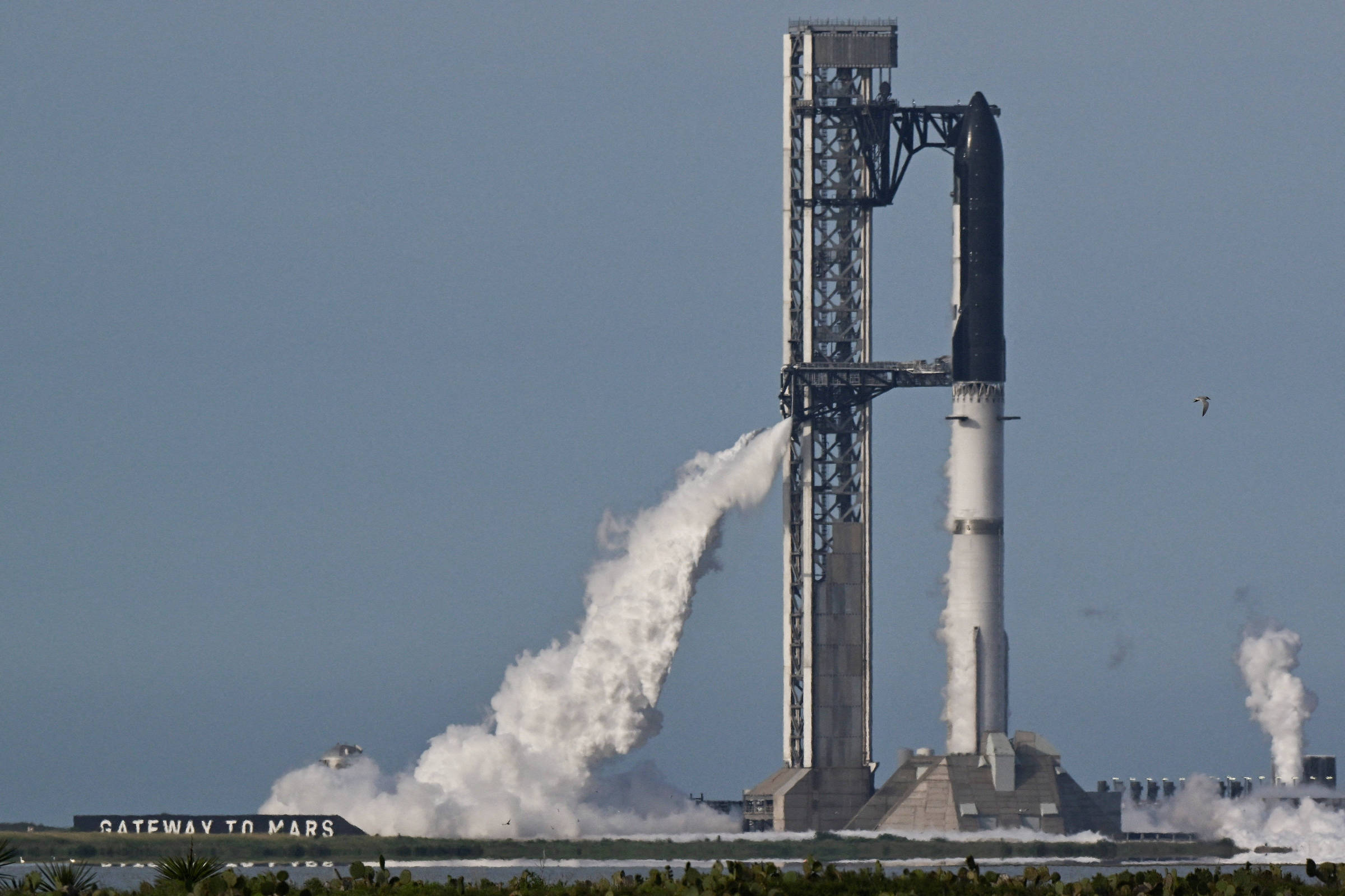 Foguete Falcon 9 da SpaceX na plataforma de lançamento com vapor saindo da base. Céu claro e vegetação ao redor da área de lançamento.