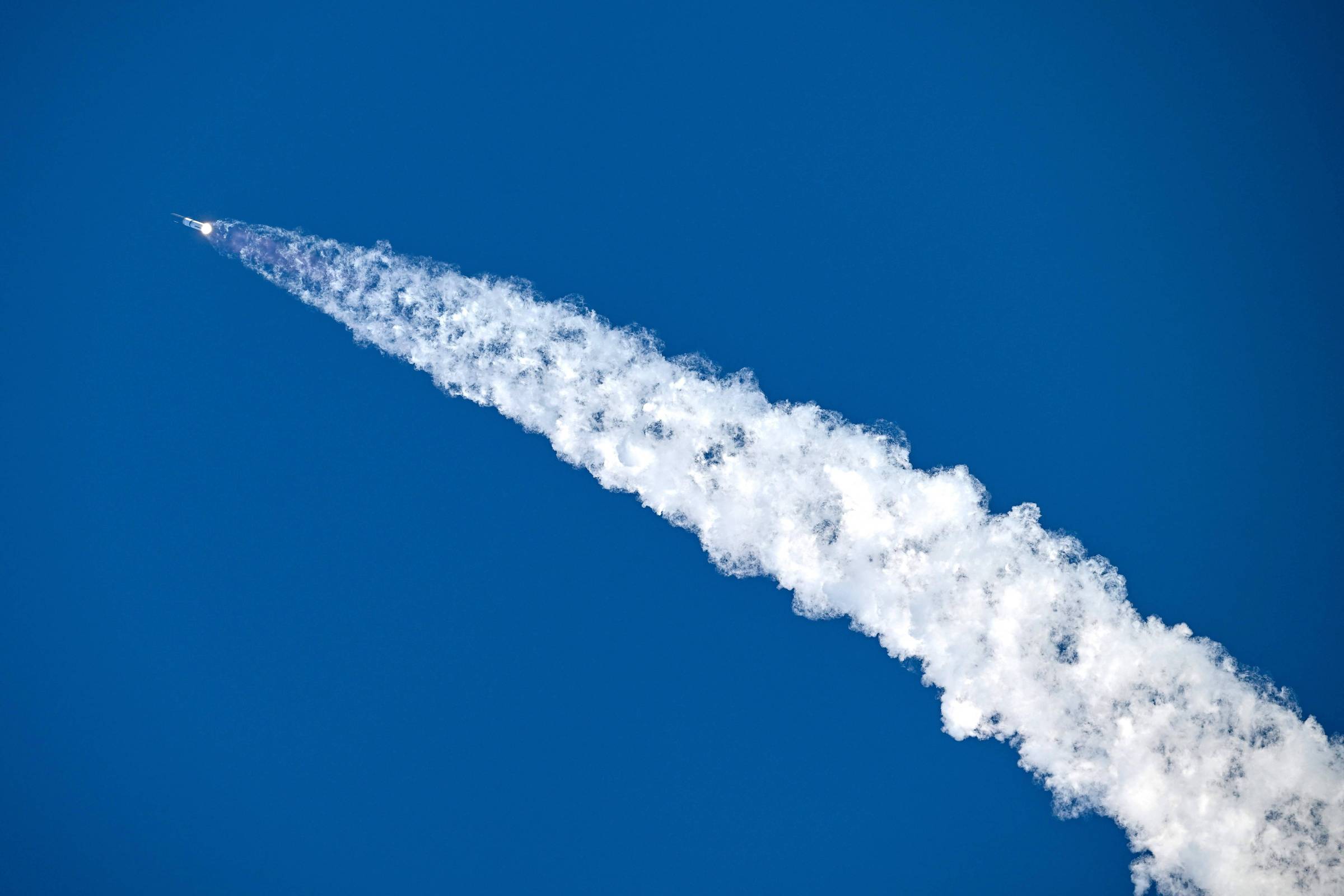 Foguete branco sobe em ângulo ascendente no céu azul claro, deixando uma longa e densa trilha de fumaça branca atrás de si.