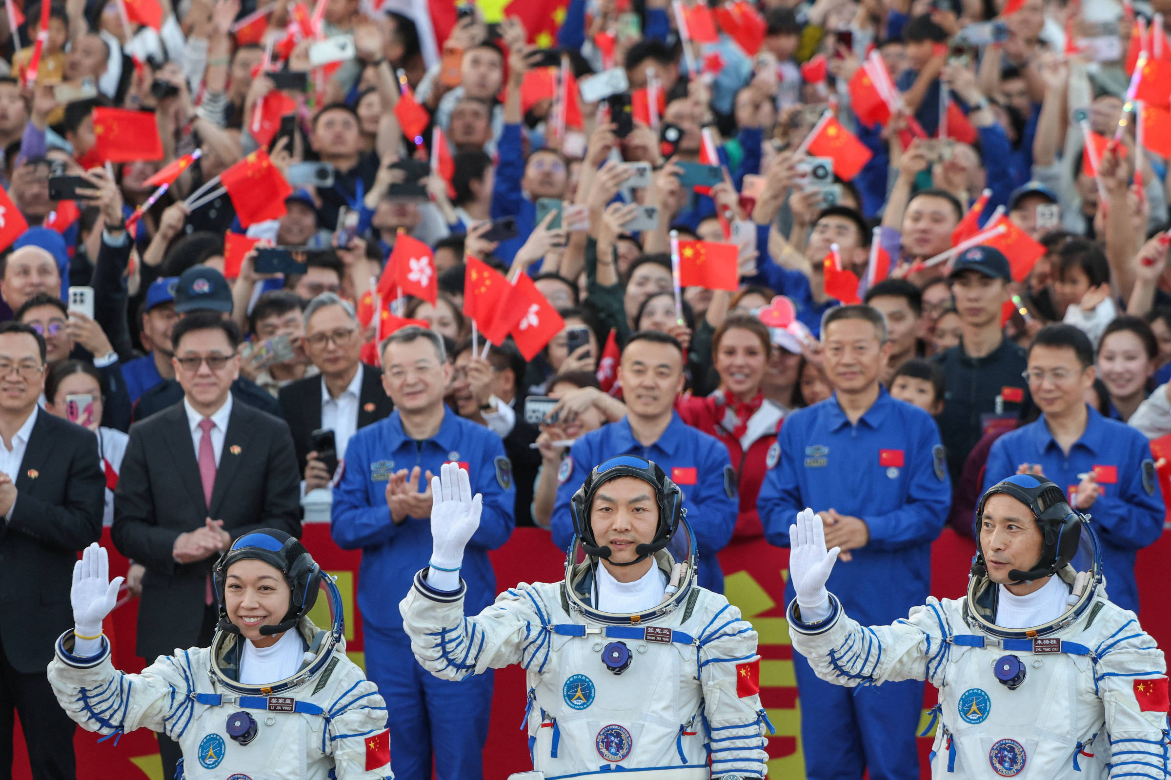 Três astronautas chineses em trajes espaciais acenam para uma multidão. Ao fundo, pessoas vestindo uniformes azuis e civis seguram pequenas bandeiras da China, celebrando o evento.