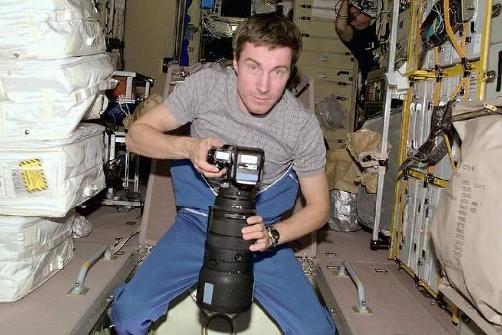 Homem com camiseta cinza e calça azul segura câmera fotográfica com lente grande, posicionado em ambiente interno com equipamentos e sacos brancos ao redor.