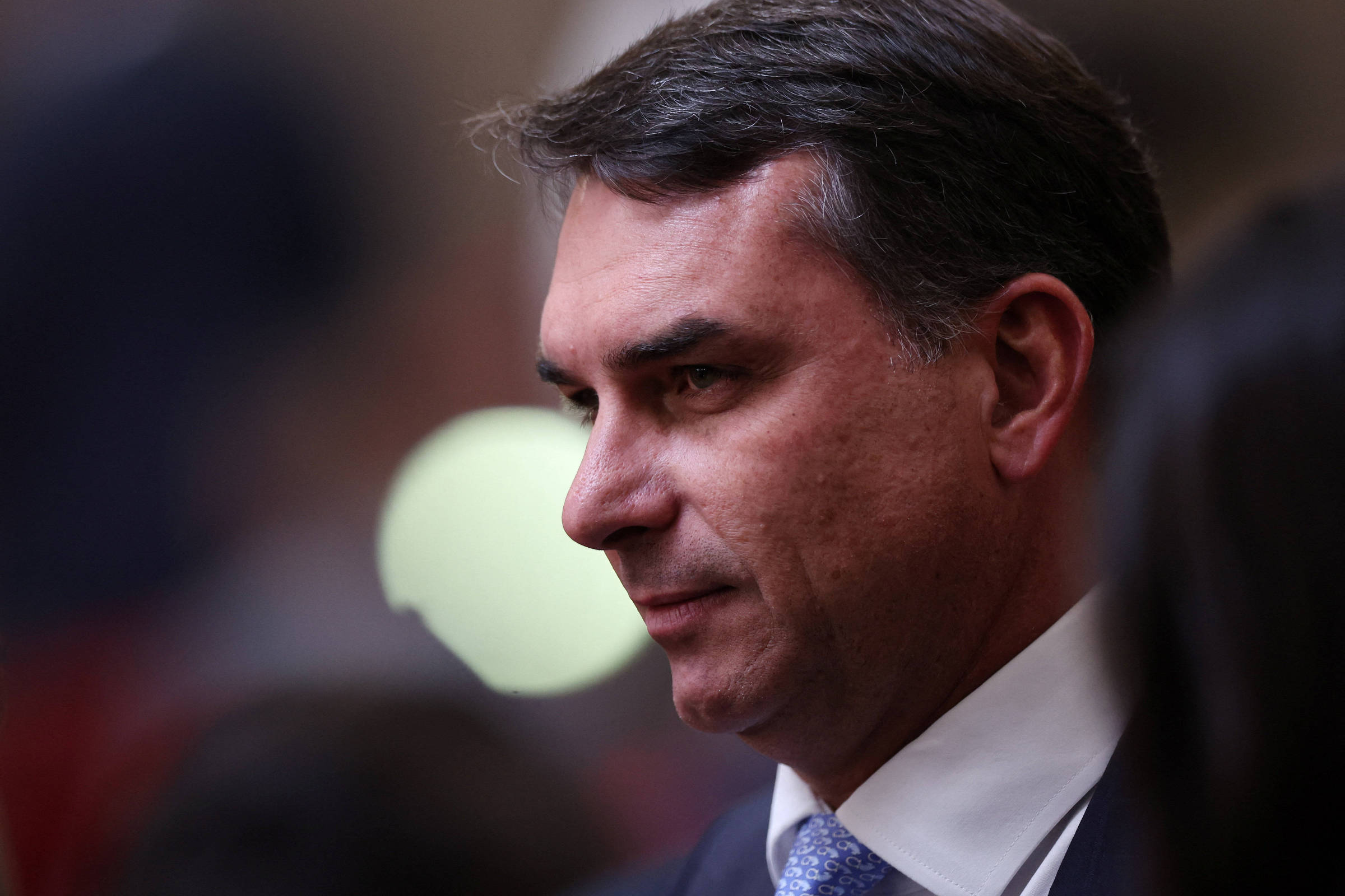 Homem de meia-idade com cabelo escuro penteado para o lado, vestindo terno escuro, camisa branca e gravata azul, em ambiente interno desfocado.