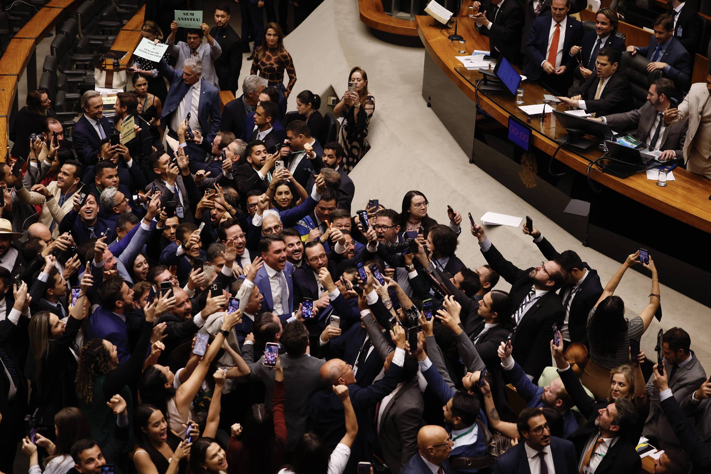 Grande grupo de deputados reunidos no plenário de uma casa legislativa, muitos tirando fotos com celulares e comemorando. Alguns parlamentares estão sentados nas mesas ao fundo, observando a movimentação.