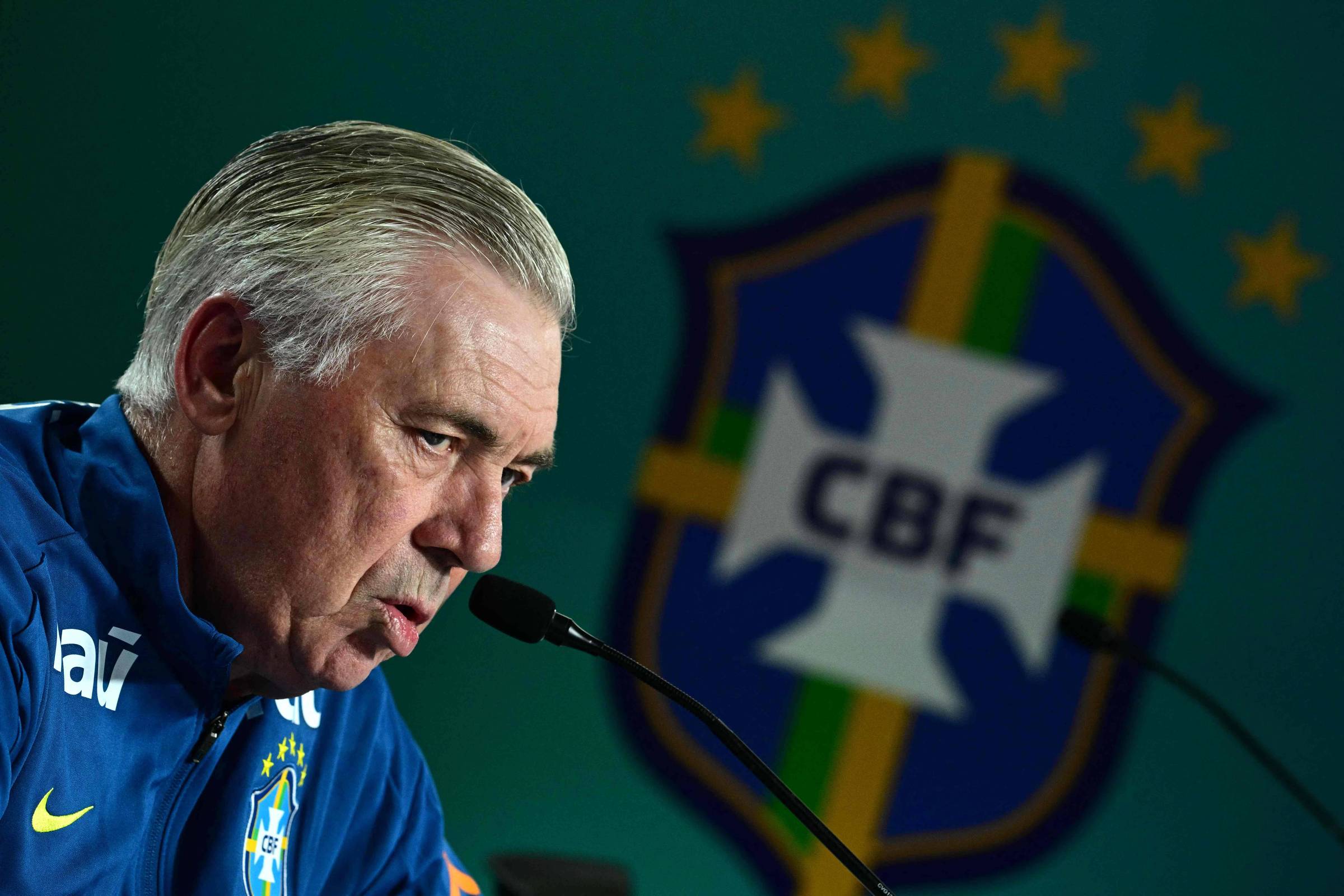 Homem de cabelos grisalhos veste agasalho azul da seleção brasileira e fala ao microfone. Ao fundo, embaçado, escudo da CBF com cinco estrelas douradas.