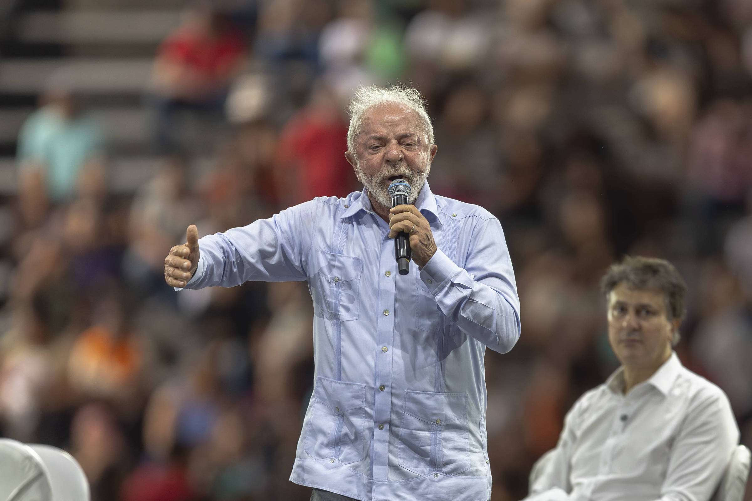 Homem de cabelos grisalhos e barba, vestindo camisa azul clara, fala ao microfone em evento com público. Duas pessoas sentadas em cadeiras brancas ao seu lado, uma delas com colete azul claro e cabelos cacheados. Plateia visível ao fundo, ambiente interno.