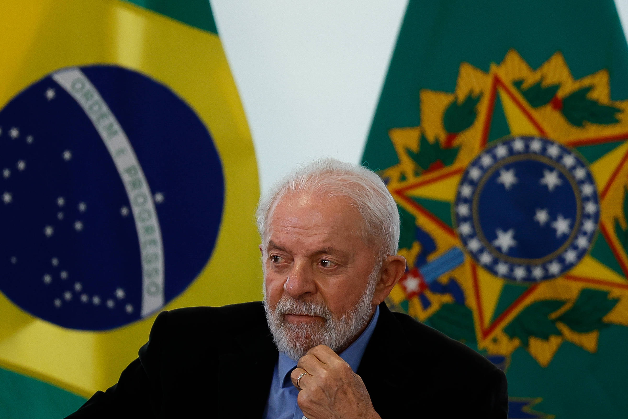 Homem de cabelos e barba brancos veste terno preto e camisa azul, sentado com mão no queixo. Ao fundo, bandeira do Brasil e símbolo nacional em fundo verde.