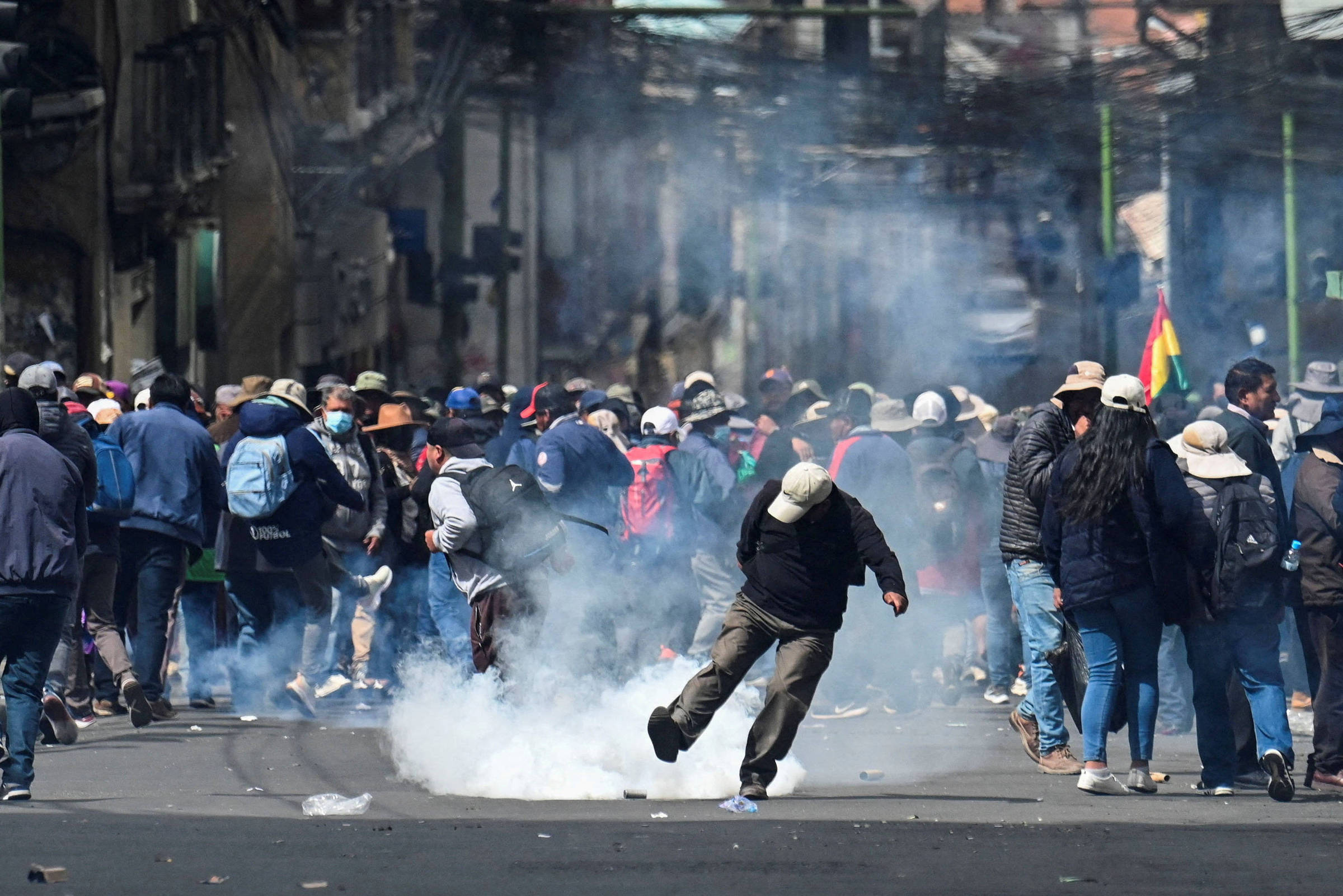 Grupo de manifestantes caminha em rua urbana enquanto um homem no centro tenta se afastar de nuvem de gás lacrimogêneo no chão. Pessoas vestem roupas casuais e chapéus, e há fumaça densa no ar.