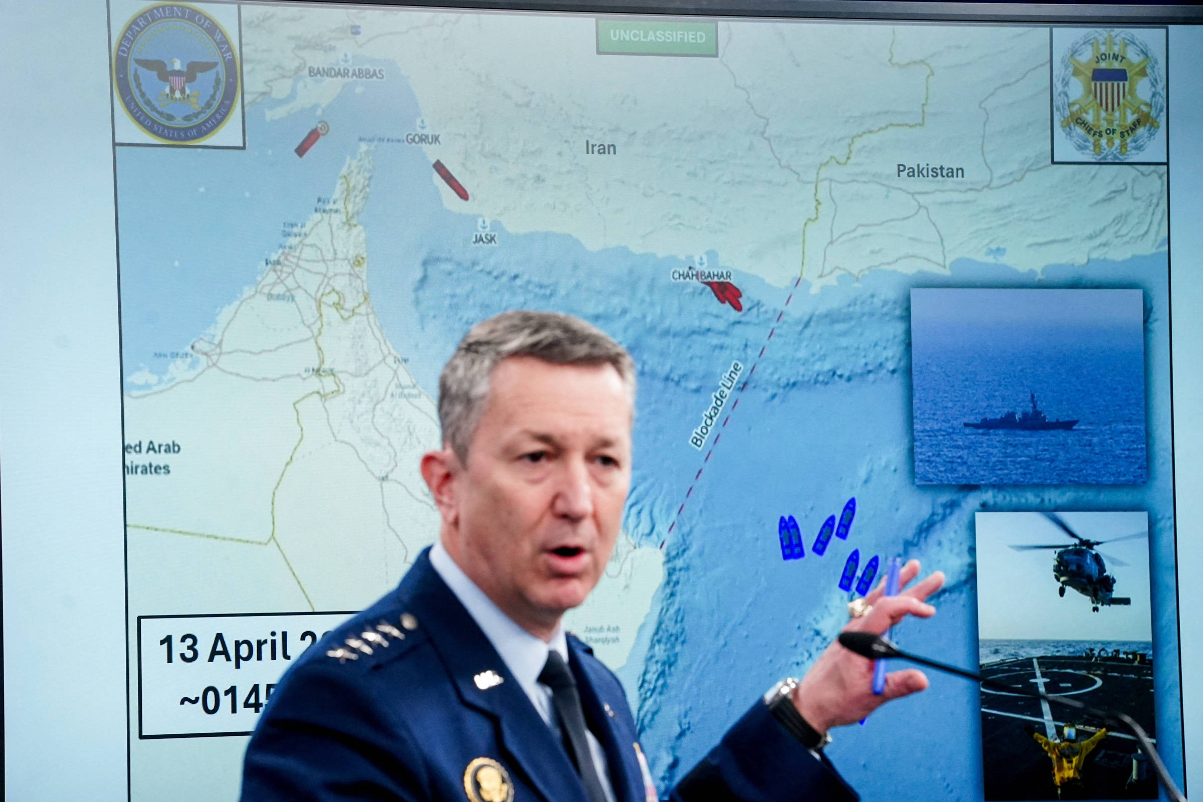 Homem em uniforme militar azul aponta para mapa do Golfo Pérsico exibido em tela, que mostra localizações de navios e aeronaves entre Irã, Arábia Saudita e Paquistão. No canto direito, imagens menores exibem um navio de guerra e um helicóptero em operação marítima.