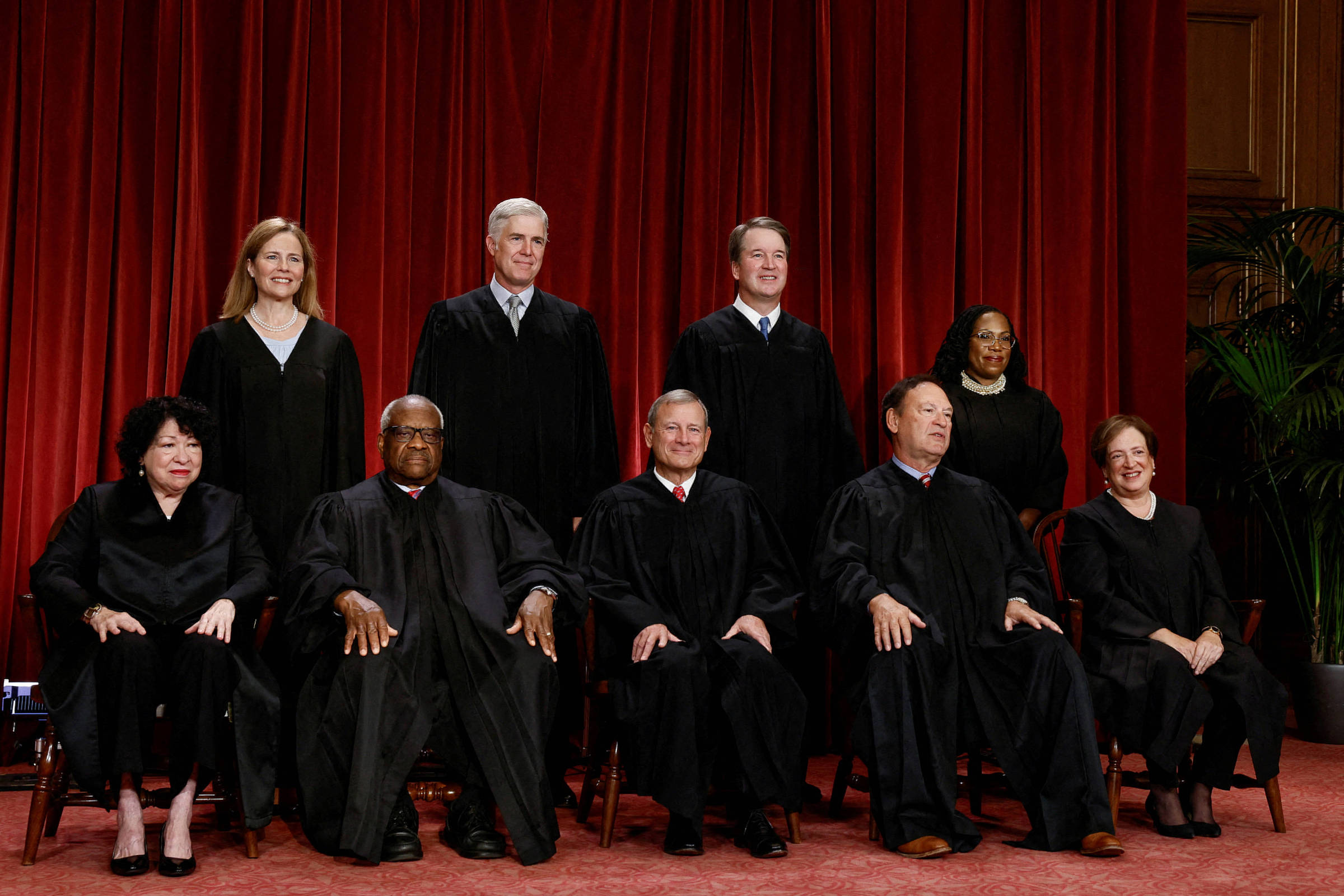 Nove juízes da Suprema Corte dos Estados Unidos posam em traje formal preto diante de uma cortina vermelha. Quatro estão sentados em cadeiras na frente e cinco em pé atrás, em ambiente interno com carpete vermelho e planta ao fundo.