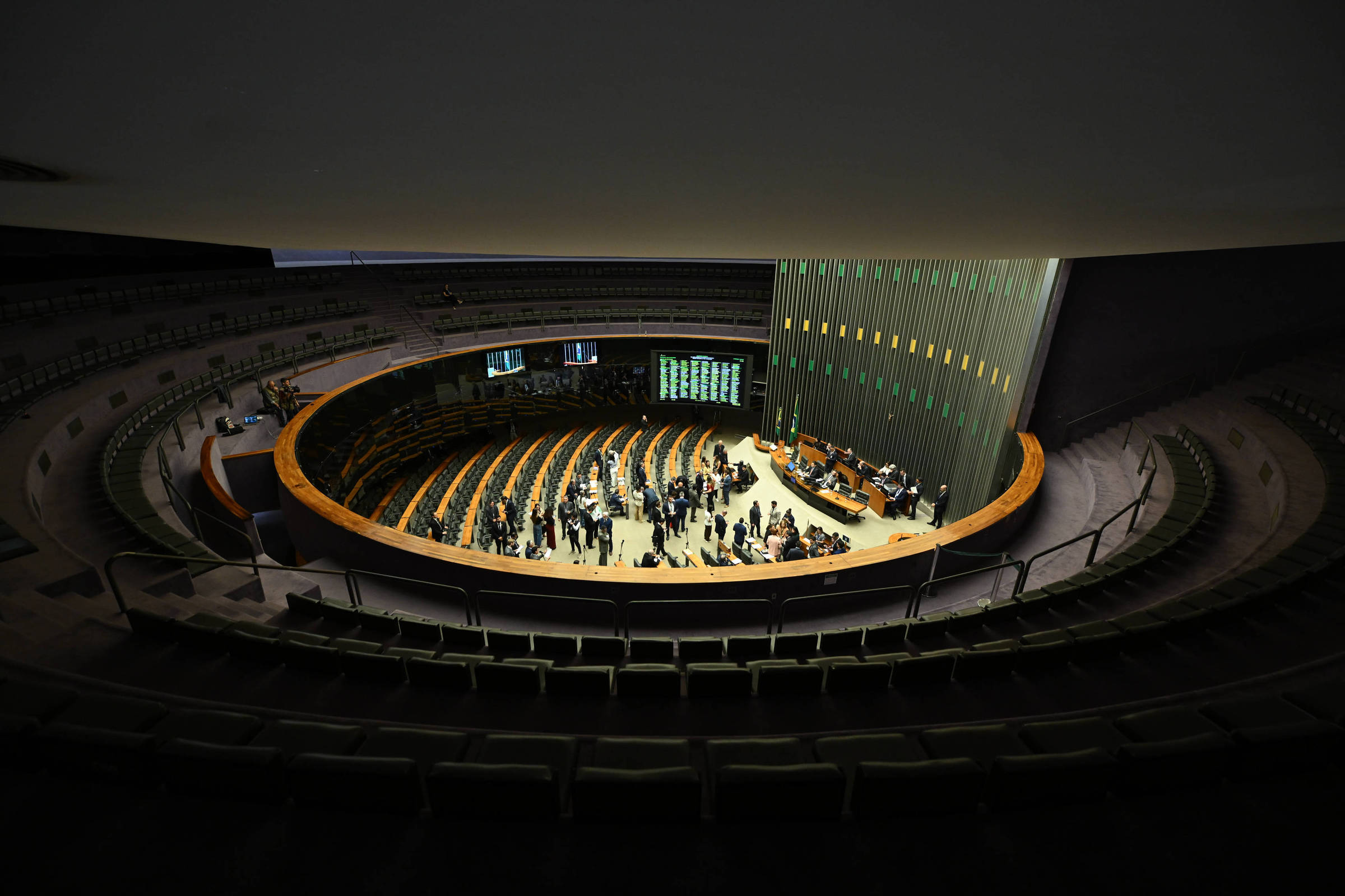 Vista ampla do plenário da Câmara dos Deputados em Brasília com deputados reunidos no centro, bancos vazios ao redor e painel eletrônico ao fundo.