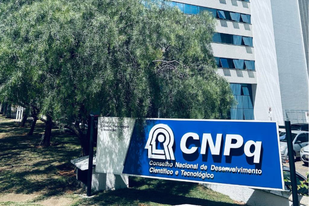 Placa azul com logotipo branco do CNPq (Conselho Nacional de Desenvolvimento Científico e Tecnológico) posicionada em área externa com árvores ao fundo e prédio alto de janelas visíveis.